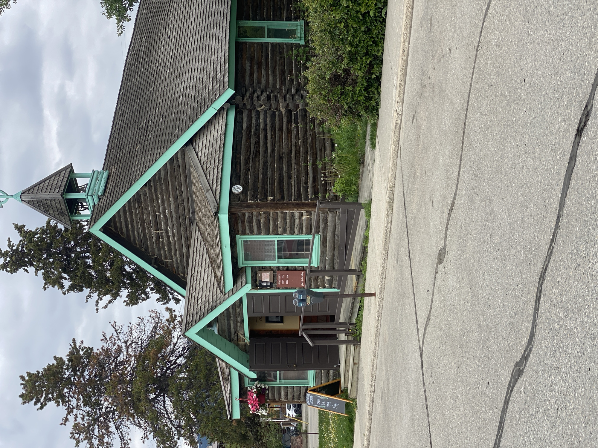 The image shows a rustic log building with a turquoise-painted roof trim and windows, possibly a historical museum, located in Whitehorse, Canada. The building has a steep gabled roof covered with wooden shingles and a small belfry with a cross on top. Double dark brown doors stand open at the entrance, flanked by windows. A ramp leads up to the entrance. To the left of the entrance, a black A-frame chalkboard sign stands on the sidewalk, displaying "WESTERN Yukon MUSEUM OPEN 10-5 DAILY" and "FREE ADMISSION". Hanging baskets with pink and white flowers are suspended above the entrance.
In front of the building, a paved sidewalk runs alongside the street. A wooden railing lines a ramp and stairs leading to the entrance. Two grey parking meters stand on the sidewalk near the entrance. A variety of green bushes and some flowering plants are visible around the base of the building. Two tall evergreen trees are behind the building. The sky is overcast with grey clouds. The street in the foreground is paved with asphalt and shows some cracks and repairs. A white car is parked on the left side of the street in the background.