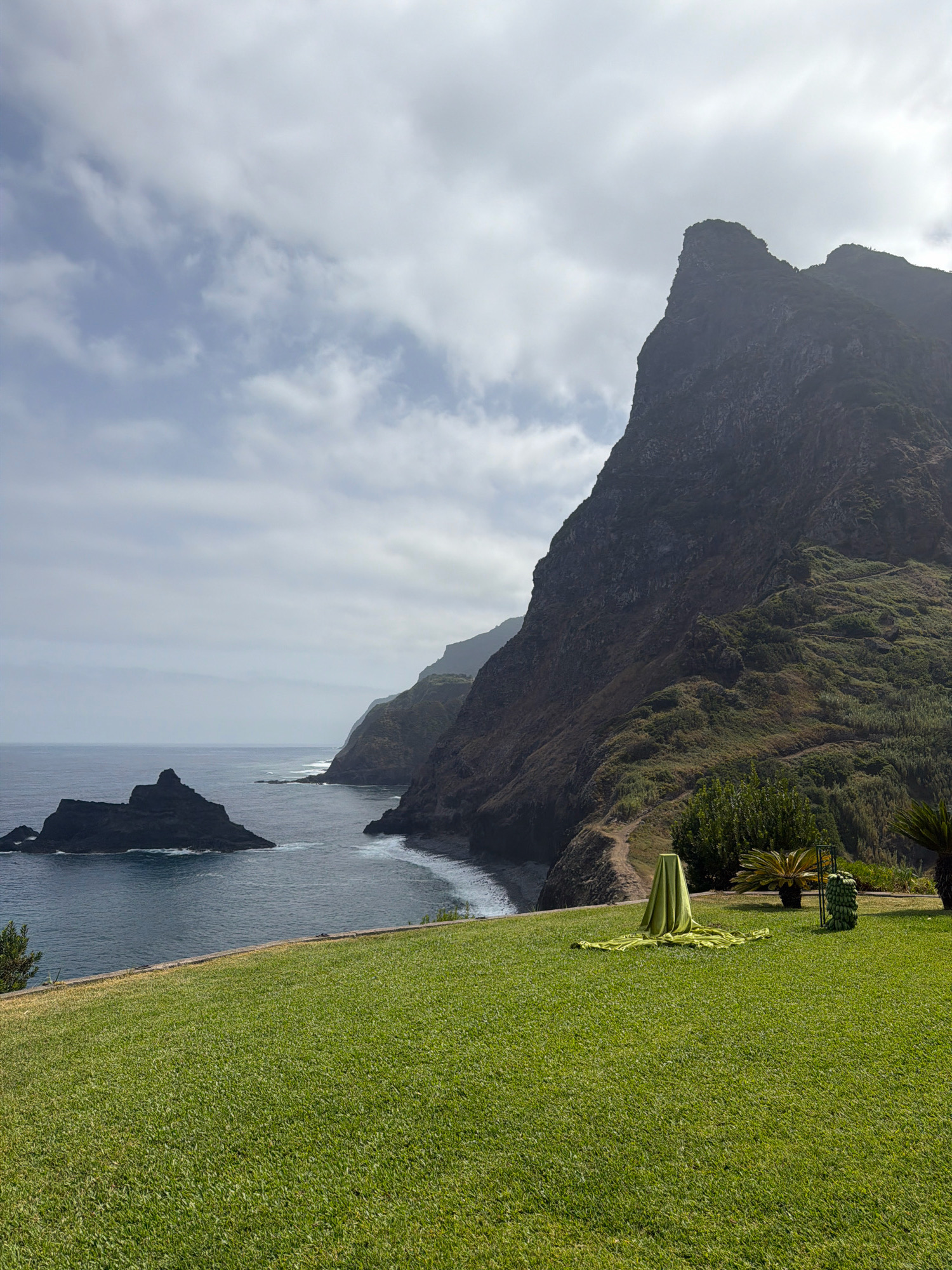 The image shows a vast, green lawn in the foreground, sloping gently upwards towards a rocky coastline. In the distance, dramatic, dark, and rugged cliffs rise steeply from the deep blue ocean, their surfaces partially covered with lush green vegetation. The sky is partly cloudy, with large, white clouds interspersed with patches of blue. On the lawn, towards the right side of the frame, a bright green cloth is draped over a triangular pedestal, and next to it, a decorative structure resembling a stack of large green objects stands. The location is the municipality of São Vicente, Portugal.
