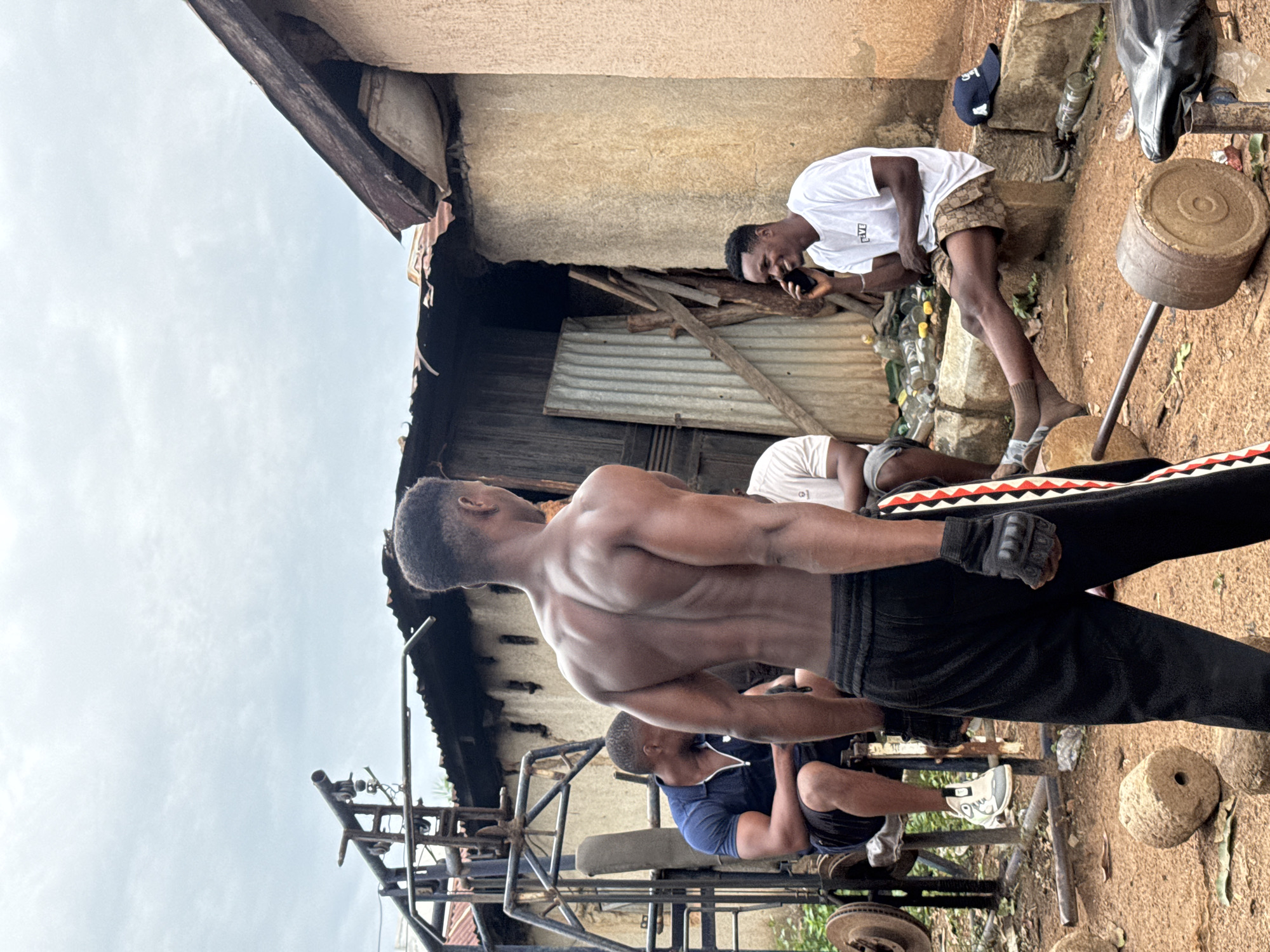In Akwanga, Nigeria, an outdoor gym is set up near a rustic, partially-constructed building with a corrugated metal roof and a textured wall. Four men are present. In the foreground, a muscular man stands with his back to the camera, wearing black workout pants with red and white stripes down the side and black weightlifting gloves. To his left, a man in a navy blue polo shirt and black shorts sits on a weightlifting bench, facing away from the camera. Another man, wearing a white t-shirt and patterned shorts, is seated on a stone block to the right, talking on a black mobile phone and smiling. A fourth man, partially visible in the background, wears a white t-shirt. The gym equipment includes a metal weight rack, weight plates, and a barbell. The ground is covered in dirt and fallen leaves, with several plastic bottles scattered around. The sky is overcast.