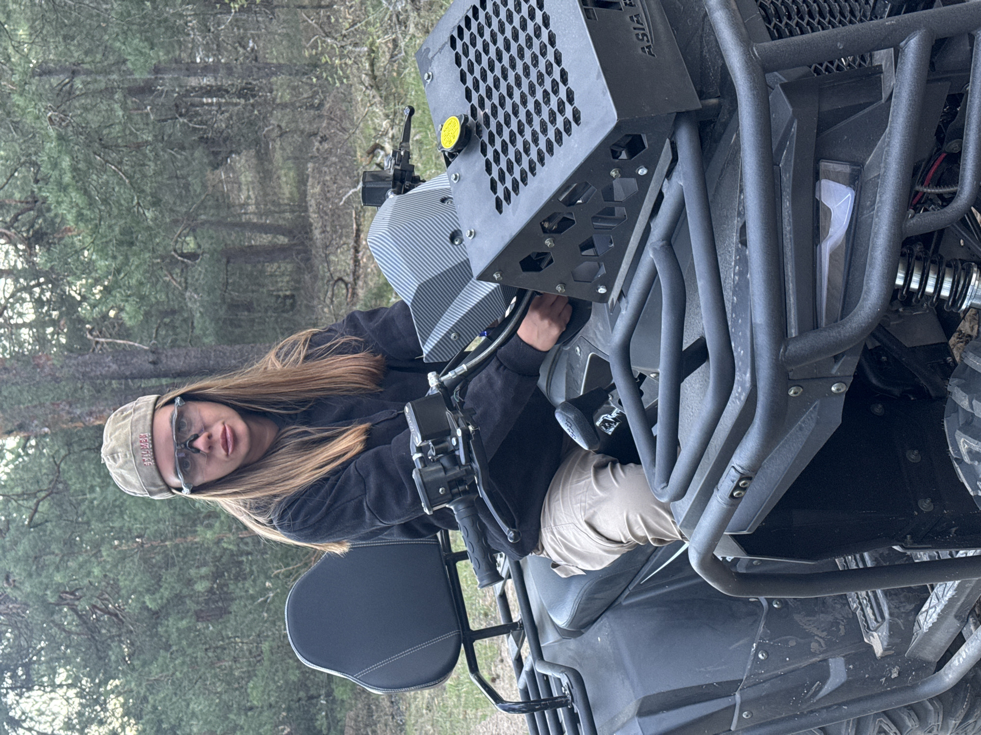 The image shows a person on a black ATV. The person is wearing a black hoodie, tan pants, glasses, and a tan cap. The ATV has a black roll cage and a radiator with a protective cage. The environment appears to be a wooded area, judging by the trees in the background. The location is Bol'shoye Kozino, Russia, as indicated by the prompt.