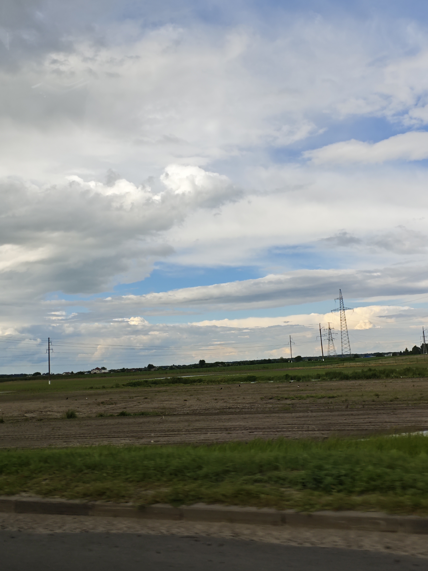 This is a medium shot of a rural landscape under a cloudy sky in the vicinity of Krasnenskoye village council, Belarus. In the foreground, a paved road with a curb runs horizontally across the bottom of the frame, partially blurred by motion. Adjacent to the road is a strip of vibrant green grass, which transitions into a brown, tilled field. In the middle ground, the field stretches towards a distant line of trees and a few buildings, some of which appear to be houses. Several power lines are visible, supported by tall, slender poles, with two larger transmission towers in the right half of the frame. The sky above is filled with a mix of light and dark grey clouds interspersed with patches of blue sky. A faint, almost imperceptible rainbow can be seen in the distance on the left side of the image. The scene is peaceful and conveys a sense of open space.