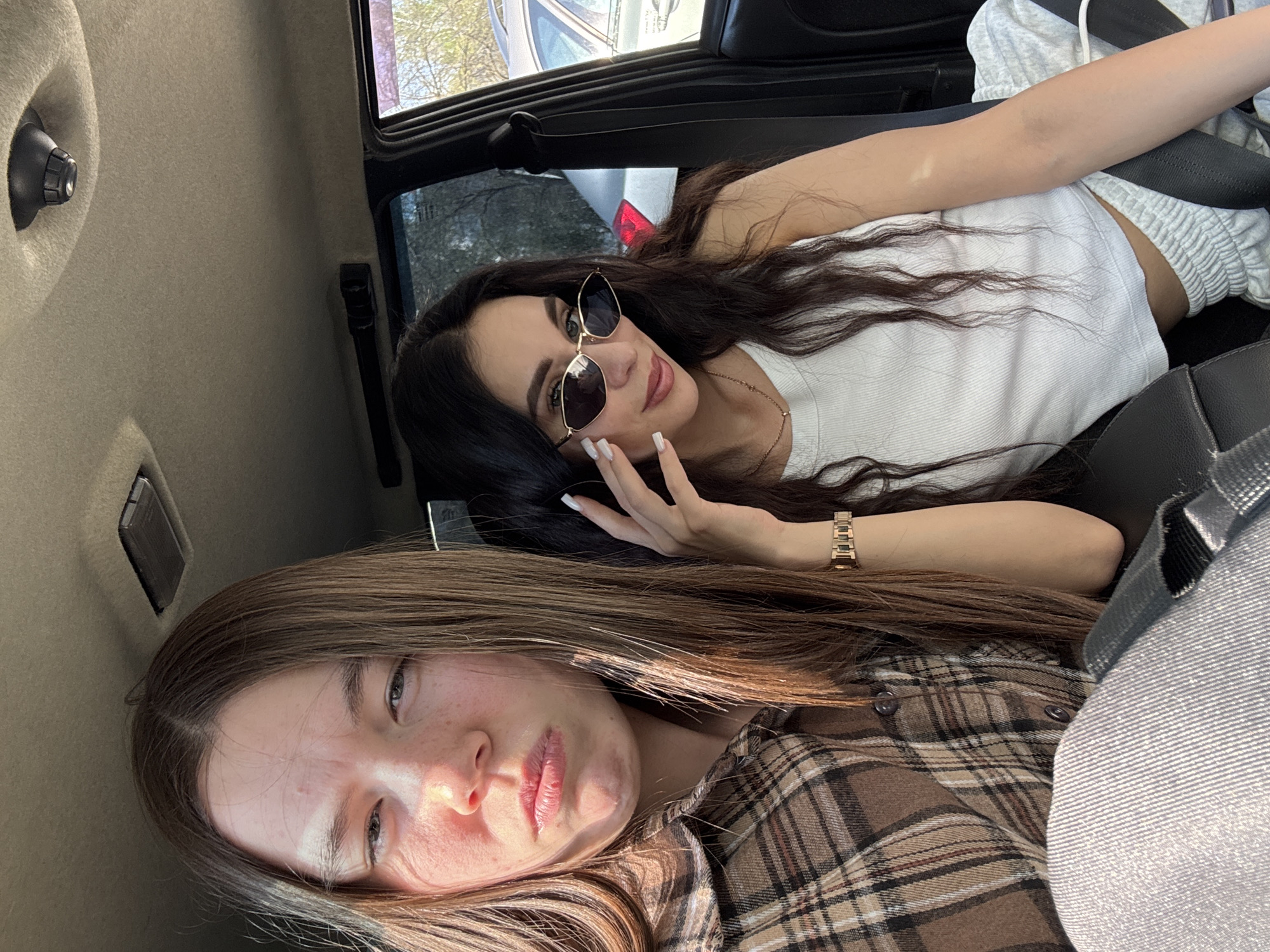 Two young women are inside a car. One woman is reclining in the back seat wearing sunglasses, a white tank top, and grey sweatpants with a seatbelt. She has long dark hair and wears a gold watch on her wrist. The other woman is reclining below her, wearing a brown plaid shirt. The interior of the car is visible, with beige panels and dark upholstery. Outside the car window, trees and other cars are visible. The image does not contain enough information to confirm the location within the city of Chaikovsky, Russia.