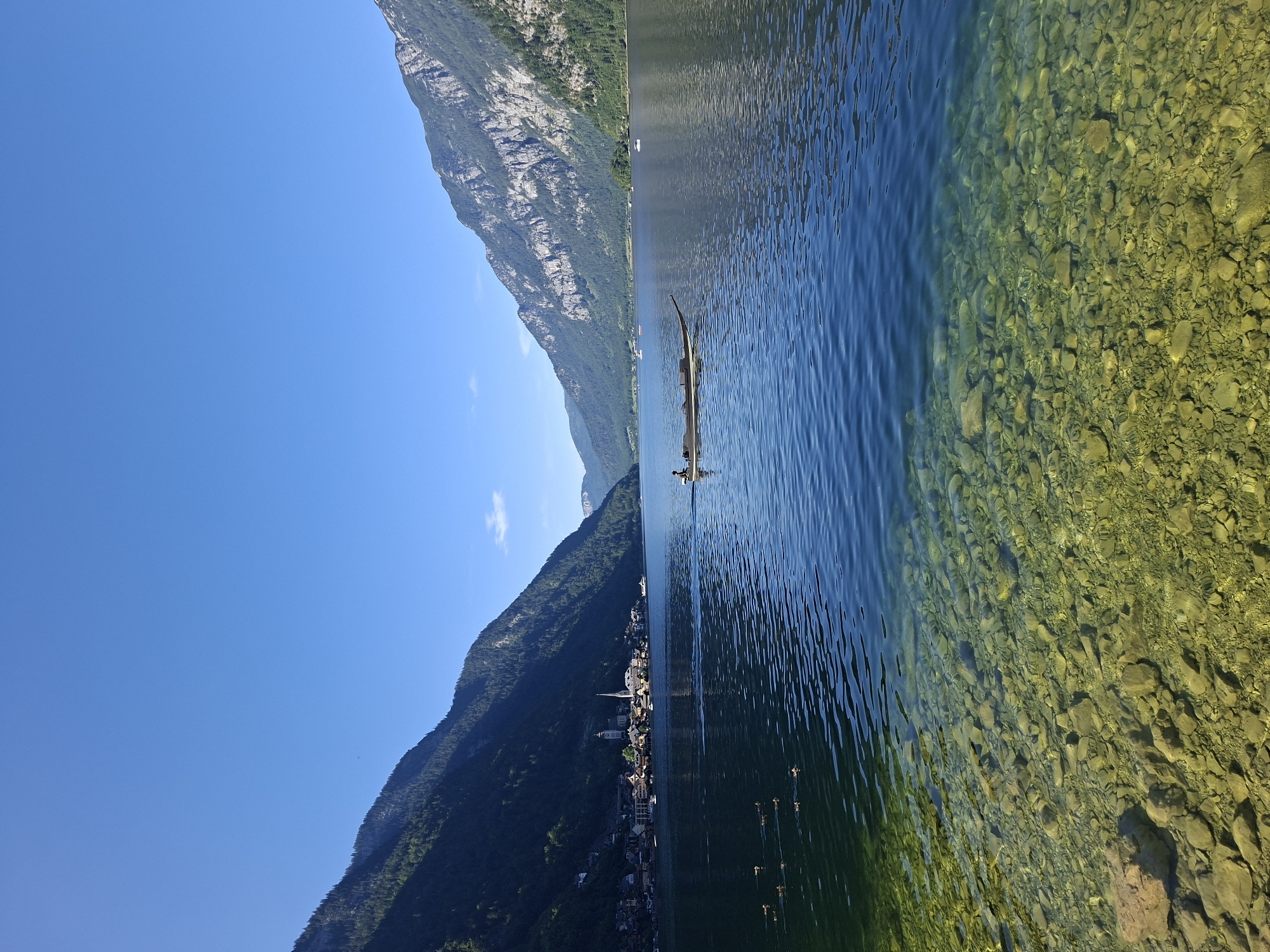 The image depicts a tranquil scene of Hallstatt Lake in Hallstatt, Austria, under a clear blue sky. In the foreground, the shallow, crystal-clear water reveals a rocky bed composed of numerous smooth, rounded stones in shades of green, yellow, and brown. The water transitions to a deeper, darker blue further from the shore. A small group of ducks swims on the surface in the middle-left of the frame.
In the mid-ground, a long, narrow wooden boat, possibly a punt, navigates the lake. A single person, silhouetted against the bright water, is seated in the boat, seemingly steering or propelling it. Further in the distance, the picturesque town of Hallstatt is visible, nestled along the shoreline. Buildings with red-tiled roofs and traditional architecture, including church spires, are clearly identifiable against the backdrop of steep, tree-covered mountains that rise dramatically on either side of the lake. The mountains are characterized by rugged, rocky outcrops and dense green foliage, with shadows cast across their slopes, indicating the time of day. Other small boats are scattered across the wider expanse of the lake.