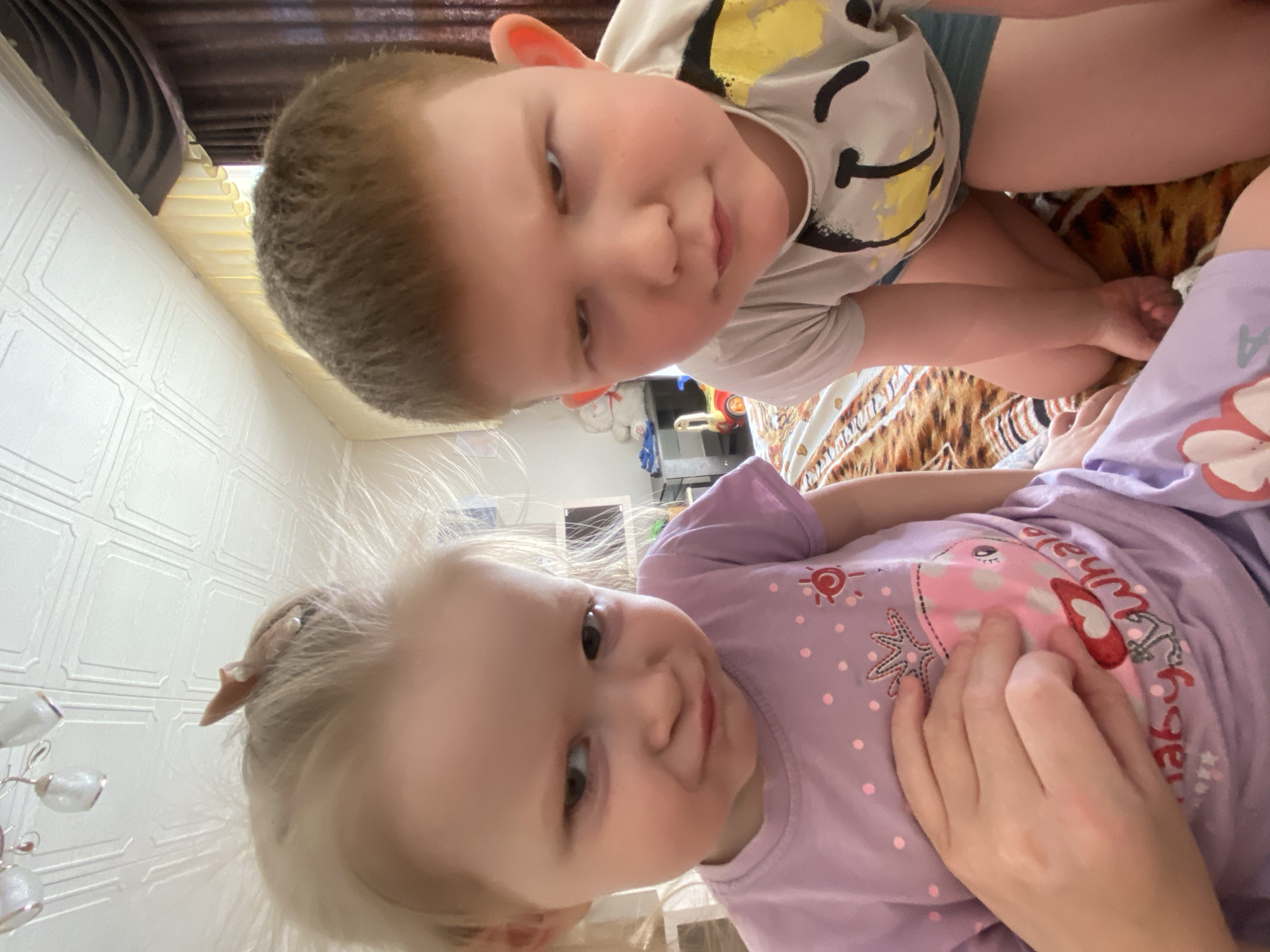 Two children are depicted in a photograph. A fair-skinned young boy with light brown hair and wearing a light-colored shirt with a smiley face design is positioned at the top of the frame. Below him is a fair-skinned young girl with blonde hair, wearing a purple t-shirt with a cartoon whale design. The girl’s hand is visible, touching the whale on her shirt. The background includes a wall and what appears to be bedding with a patterned design. The photo was taken in an indoor setting in the city of Chaikovsky, Russia.