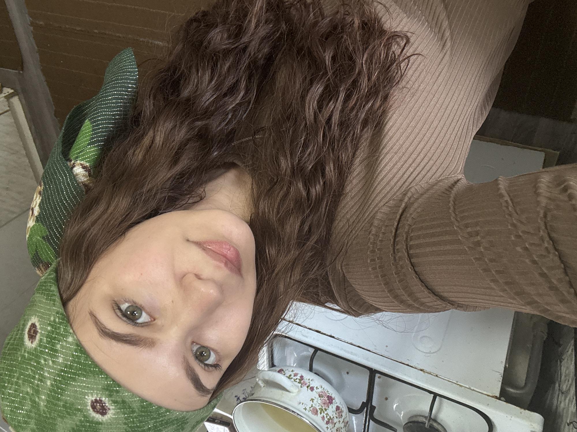 A young woman with long, curly brown hair and green eyes is the subject of a selfie. She wears a green headscarf with a subtle pattern. She is wearing a brown, ribbed top. The photo appears to be taken in a kitchen; a white stove with a pot on it is visible. Location is unspecified.