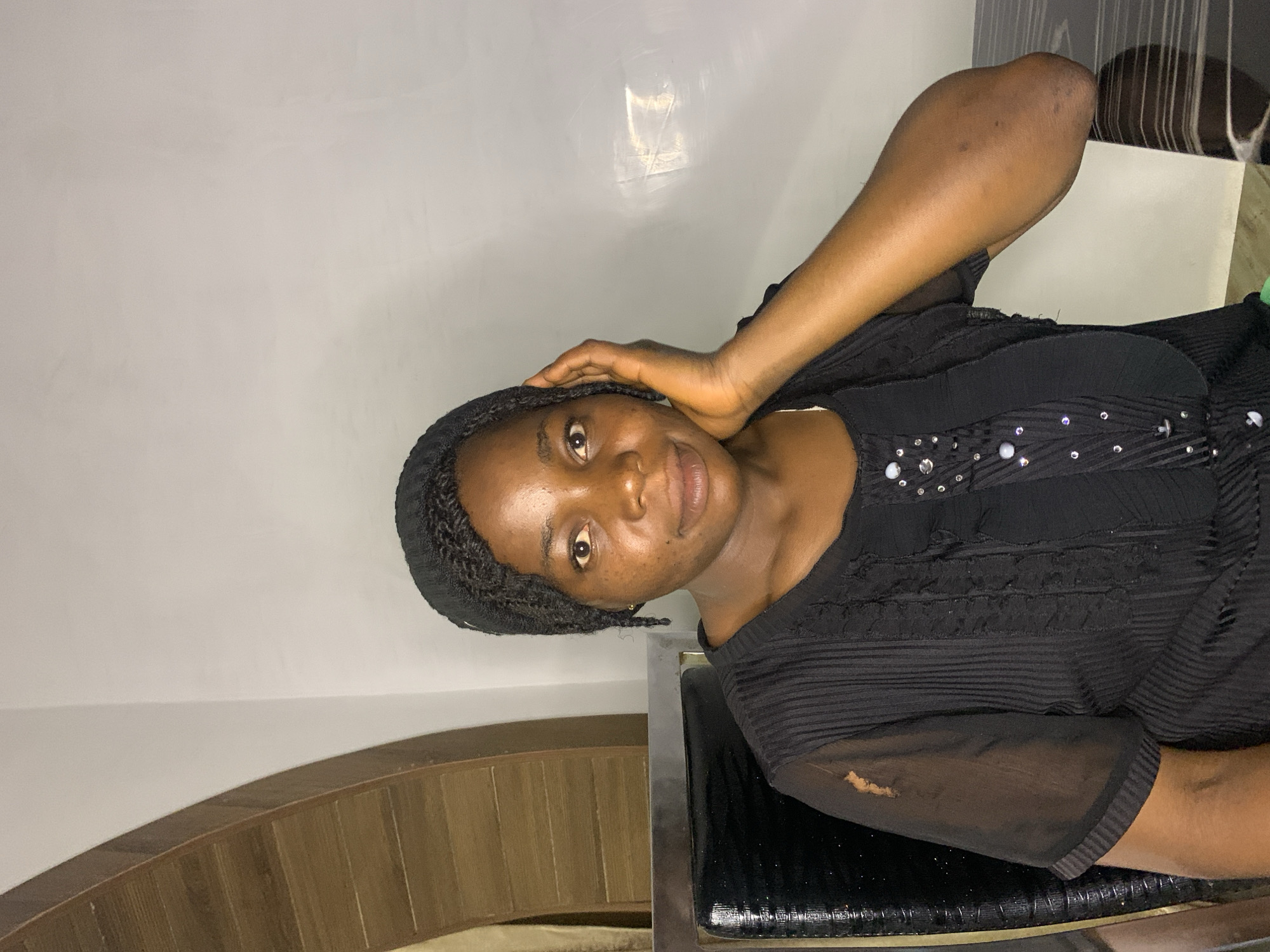A medium shot captures a woman with dark skin and braided hair wearing a black top. She is seated in a chair, with her right hand resting on her cheek. The background features a white wall and a wooden architectural element on the left. The scene is indoors, possibly in a home or office setting in Jos, Nigeria.