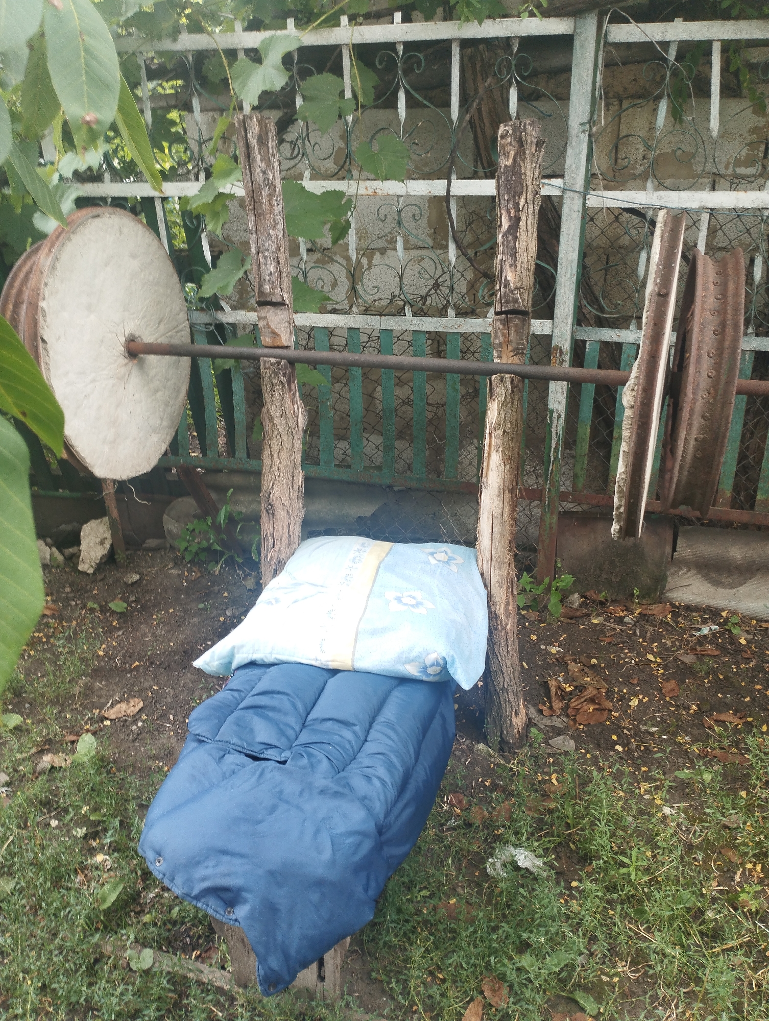 The image shows a makeshift weightlifting bench set up outdoors in Ciuciulea, Moldova. The bench consists of a blue sleeping bag and a pillow placed on a wooden surface. A barbell is supported by two rough wooden posts. On the left side of the barbell, a large, round, pale gray weight, possibly made from concrete or stone, is attached. On the right side, two heavy, irregularly shaped rusty metal objects, likely weights or scrap metal, are also attached to the barbell. The background includes a metal fence with a decorative pattern, some greenery, and what appears to be a stone wall or structure. The ground is covered with dirt, grass, and fallen leaves.