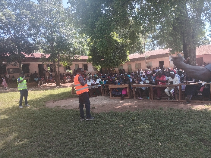 An outdoor daytime scene depicts a public gathering in Idakho South ward, Kenya. In the foreground, a grassy area transitions into a dirt path. Two men are standing; one on the left wears a yellow-green reflective vest and dark pants, while another in the center wears an orange reflective vest, a patterned shirt, and dark pants, appearing to address the assembly with his hand raised near his face. A large crowd, estimated to be over 100 individuals, primarily adults, is seated on multiple rows of rustic wooden benches facing the two standing men. Several individuals within the seated crowd are observed clapping. In the background, beneath mature trees, single-story buildings with light-colored walls and reddish-brown roofs are visible, with additional people gathered in front of them. The environment is illuminated by bright sunlight, casting distinct shadows.