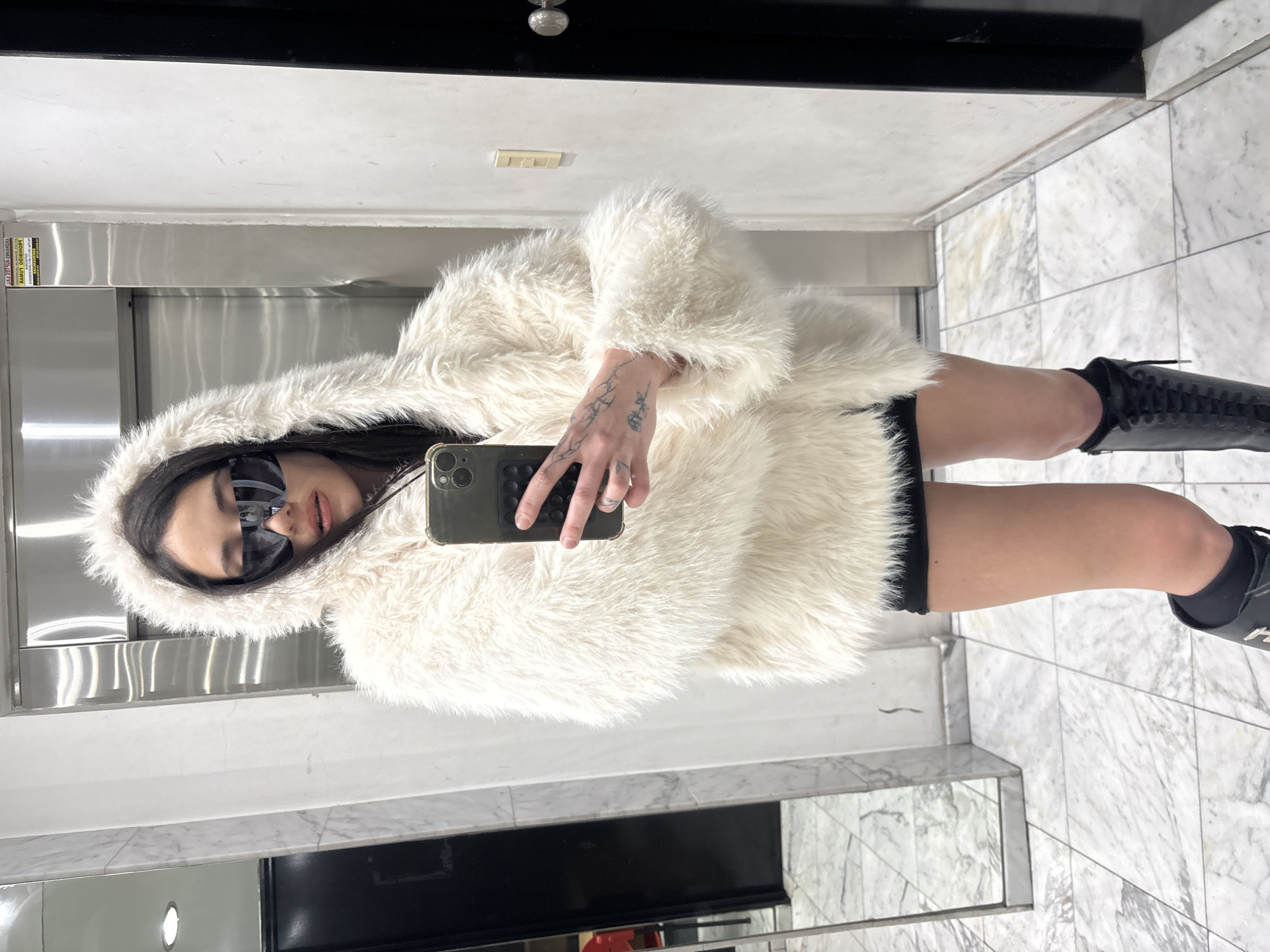 A young woman takes a selfie in a mirrored elevator. She is wearing a fluffy white hooded coat, sunglasses, a black skirt, and black boots. The elevator interior is modern, with stainless steel panels and marble flooring. The location is likely a hotel or apartment building. No visible details indicate it is Buenos Aires, Argentina.