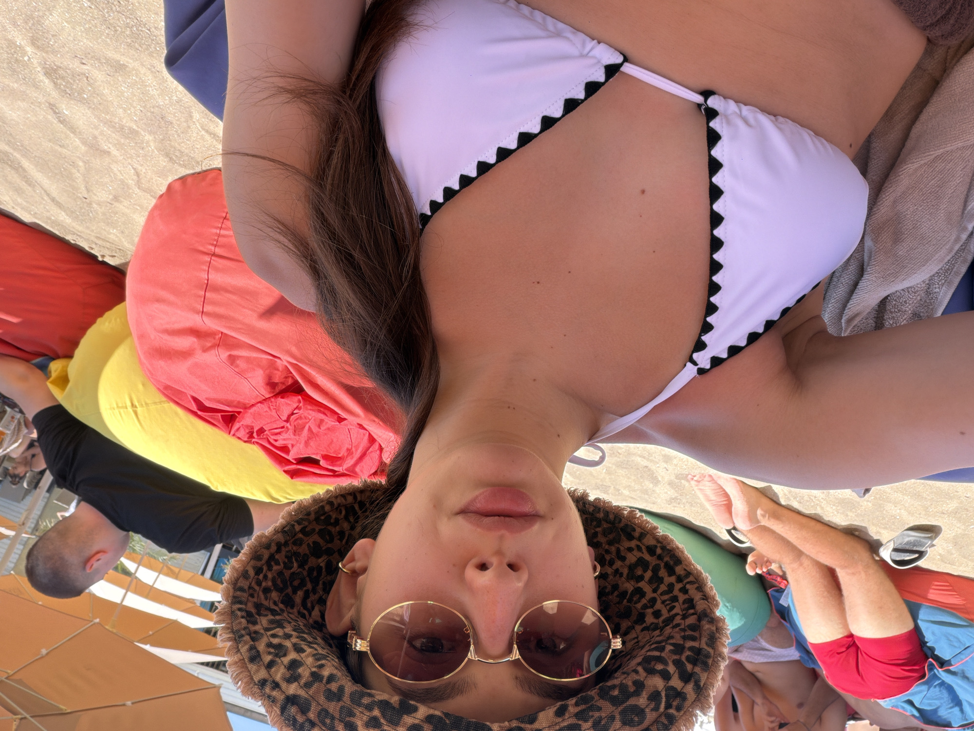 A young woman with brown hair, wearing a leopard-print bucket hat and round sunglasses, is taking a selfie on a beach. She is wearing a white bikini top with black scalloped trim. Her right arm is resting on her chest. In the background, people are seen sitting on beanbag chairs and umbrellas are visible. The environment is a sandy beach. The scene is set in Sozopol, Bulgaria, specifically on the beach.