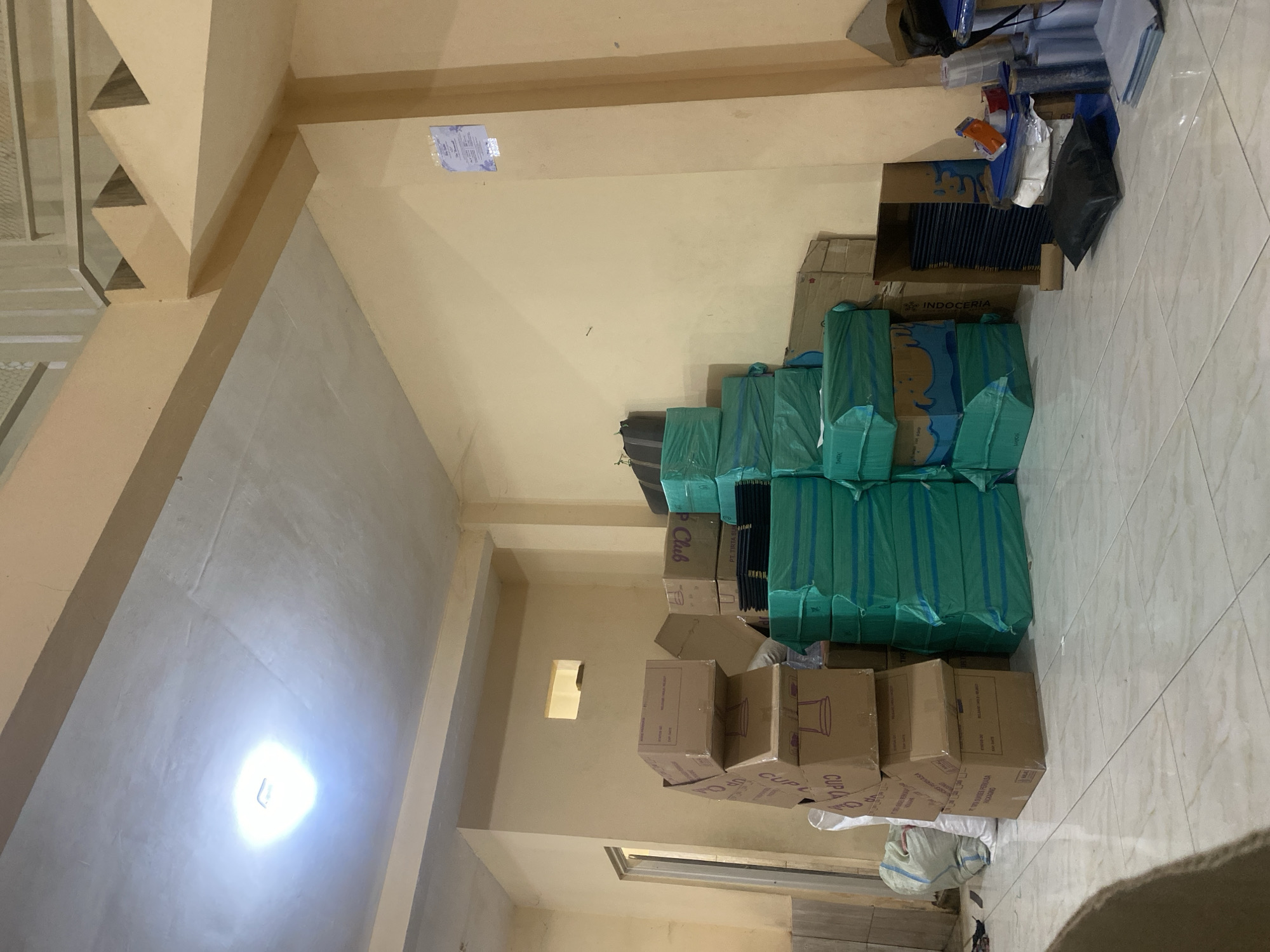 This is a medium-angle, eye-level shot of a storage room in a commercial building. The walls and ceiling are painted a light beige, and the floor is tiled with a light gray and white pattern. A bright LED light is visible on the ceiling. The room is filled with stacks of boxes and other items, suggesting a storage or warehouse environment.
In the left portion of the image, a stack of brown cardboard boxes is piled high, with some boxes labeled "CUP." To the left of these boxes, a large white woven bag is partially visible.
In the center of the image, a large stack of rectangular items wrapped in teal plastic is dominant. These appear to be uniformly sized and are neatly stacked. Adjacent to these teal-wrapped items, to the right, is a stack of brown cardboard boxes, some with text visible, including "PT. TIRTA SU..." and "Club."
Further to the right, a tall brown cardboard box stands open, revealing stacks of dark blue, rectangular items inside. In front of this open box, a black pillow or cushion lies on the floor. Next to the open box and behind the teal-wrapped items, are various other objects, including stacks of plastic containers, rolls of clear plastic wrap, and other boxes.
A staircase with beige risers and treads is visible in the upper right section of the image, leading upwards. A notice or poster is affixed to the wall on the right side of the room. The overall impression is one of organized but extensive storage of goods, likely for distribution or sale.
The image is dated October 12, 2023, at 12:43 AM, and was geotagged to Karangbong, Indonesia. The conditions appear to be interior, artificial lighting, and a stable, dry environment. There are no people visible in the scene.
