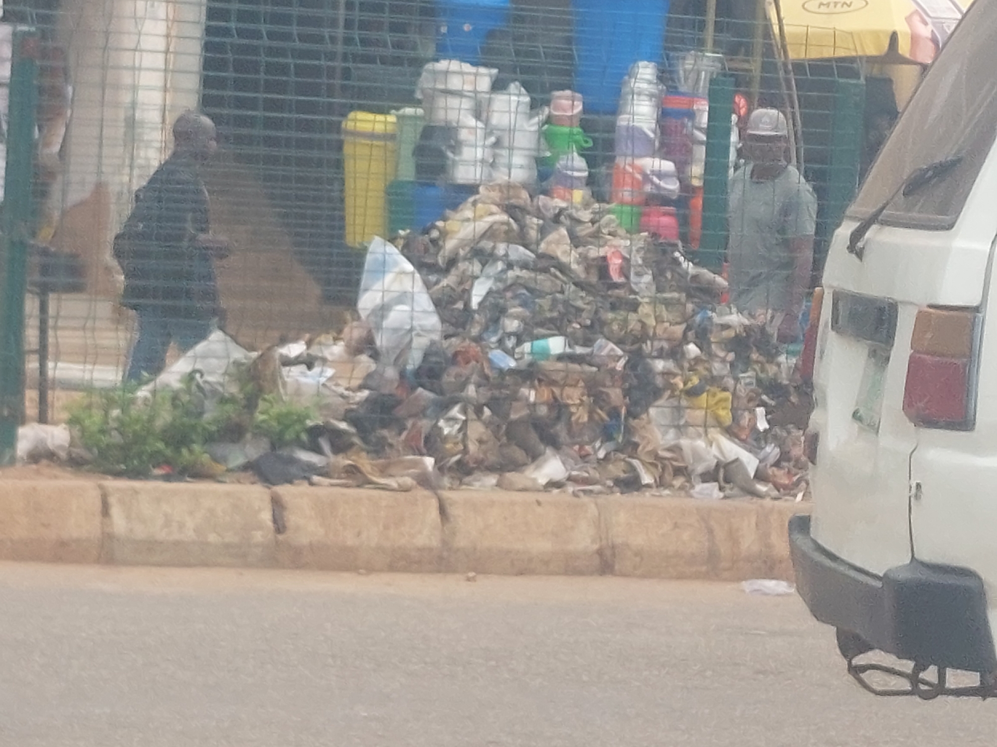 A pile of trash is visible on the side of a road in an urban environment. Two men are present. One man, wearing a dark jacket and blue jeans, walks away from the viewer and the trash pile. Another man, wearing a light green t-shirt and a white cap, stands facing the trash pile. In the foreground, the rear of a white vehicle is visible. The scene appears to be an outdoor market or street vendor area, with a mesh fence behind the trash pile and a yellow awning visible in the background. The ground in front of the trash pile is a concrete curb, and the road is paved.