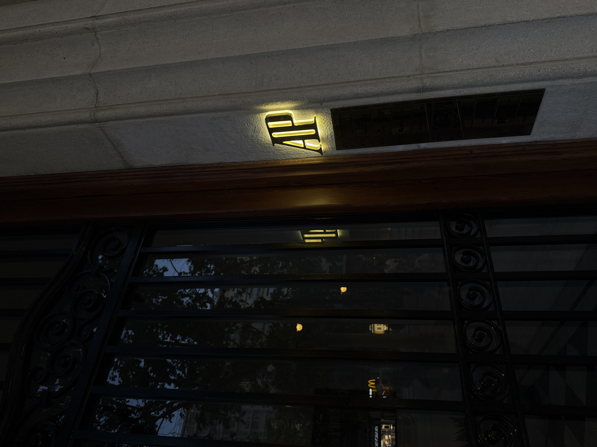 The image depicts the exterior of a building in Barcelona, Spain, captured at night or late dusk. A prominent, gold-colored "AP" sign, illuminated with a warm yellow glow, is affixed to a textured, light-grey stone facade. Adjacent to the "AP" sign on the right is a dark, rectangular plaque with several lines of faint, almost illegible text. Upon close inspection, some words or fragments such as "4°C", "AUTO CROP", "SYSTEM", "NYLONS", "GROUP", and "PARK" can be faintly discerned, suggesting a directory or list of tenants.
Below the sign and plaque, a dark wooden beam or molding extends horizontally across the frame. Beneath this, a large window or glass entrance features decorative black wrought-iron grillwork with scroll patterns. The glass reflects elements of the urban environment, including the dark evening sky, tree foliage, and distant building outlines. Several bright, yellow light sources are reflected within the glass, along with a distinct "M" logo, characteristic of McDonald's, and other blurred, inverted text from street-level signs. No people or specific activities are visible, focusing instead on the architectural details and the interplay of light and reflection in the nighttime setting.
