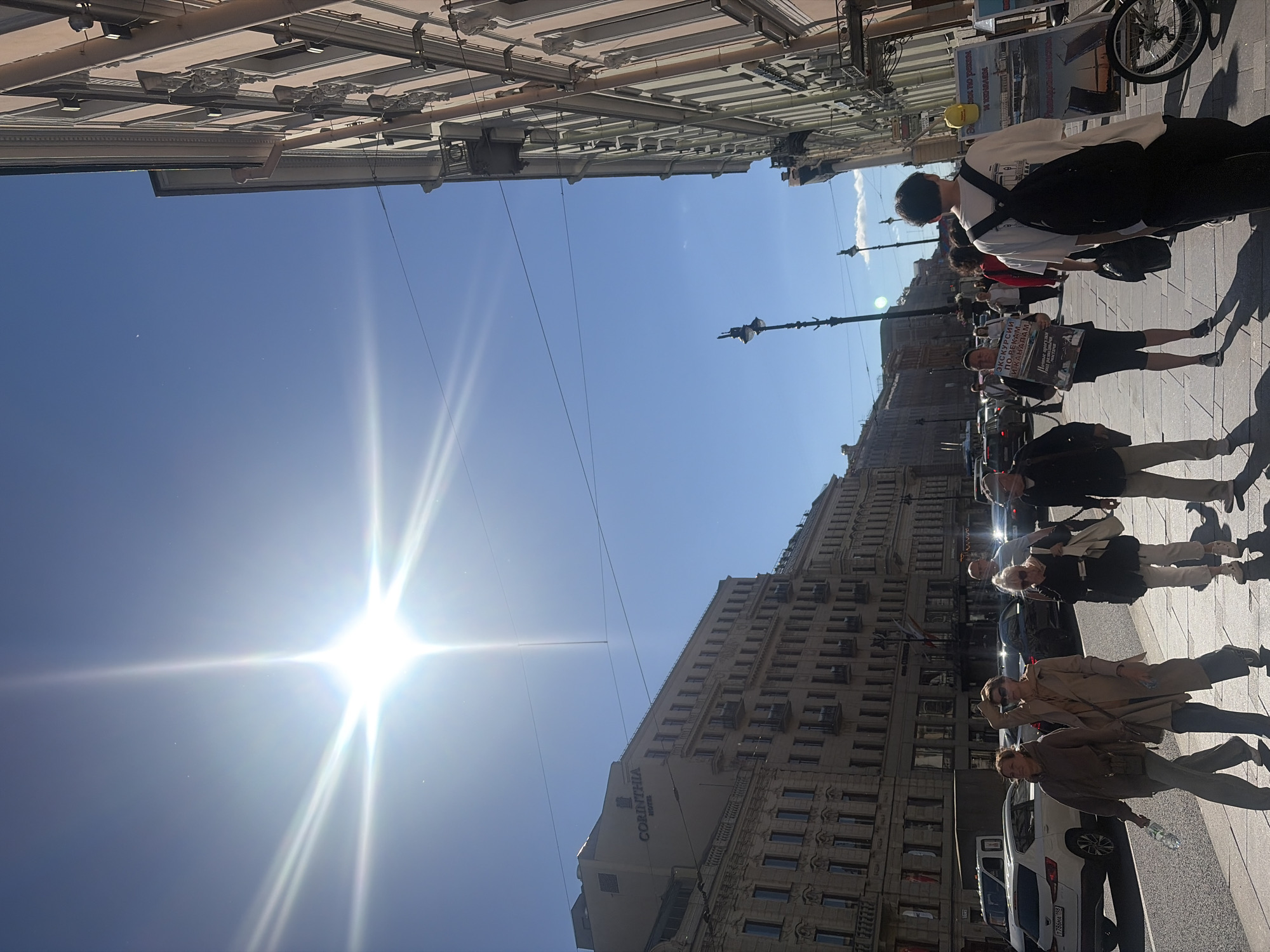 The image depicts a street scene in Saint Petersburg, Russia, on a bright, sunny day. The sun is high in the clear blue sky, creating a strong lens flare effect. On the left side of the frame, several multi-story buildings line the street, some with visible signage, including "CORINTHIA HOTEL". On the right, a similar ornate building with multiple windows and architectural details dominates the view. The street is paved with cobblestones, and a sidewalk runs along the buildings.
Several people are walking on the sidewalk. In the foreground, on the left, a woman in a brown hoodie and gray pants walks while holding a plastic bottle. Next to her, a woman in a beige trench coat and dark pants is also walking. Further down the sidewalk, a woman in a black top and white pants with a large tote bag walks, and behind her, a man in a dark jacket and hat is visible. In the middle ground, a group of people are walking, some holding newspapers or bags. One man is holding a sign that reads "ЭКСКУРСИИ ПО РЕКАМ И КАНАЛАМ" which translates to "Excursions on rivers and canals." A young person with a backpack is walking away from the camera.
Several cars are parked and driving on the street, including white vans and sedans. A bicycle is visible on the far right of the frame, leaning against a stand with advertising posters. Streetlights and overhead wires are also present. The overall impression is of a bustling urban environment under a clear sky.