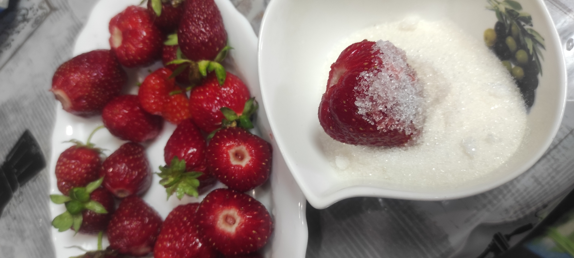 A still life photograph displays a white plate filled with fresh strawberries. In the foreground, a white bowl contains sugar, with a single strawberry partially submerged in it, its bottom half coated in sugar crystals. The scene is set on a surface that appears to be a tablecloth with a subtle checkered pattern. The image is likely captured in Kobeliachok, Ukraine.