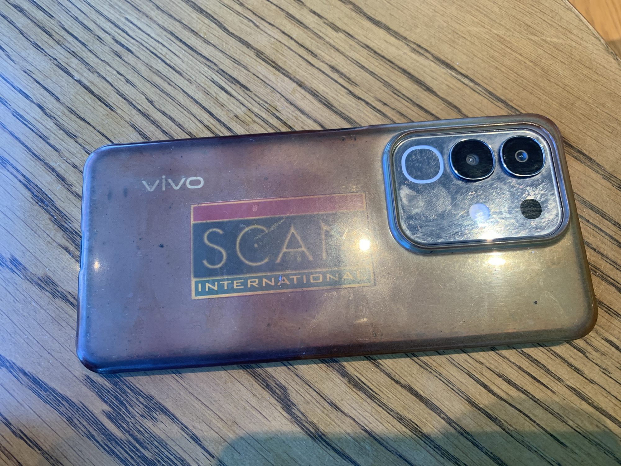 The image shows the back of a smartphone with a distinctive "SCAM INTERNATIONAL" sticker partially covering the camera array and a "VIVO" logo near the top. The phone is placed on a wooden surface with a prominent grain pattern. The lighting suggests it is daytime, casting shadows and highlights on the phone and the table. No specific location in Angeles City, Philippines, can be determined from the image alone.