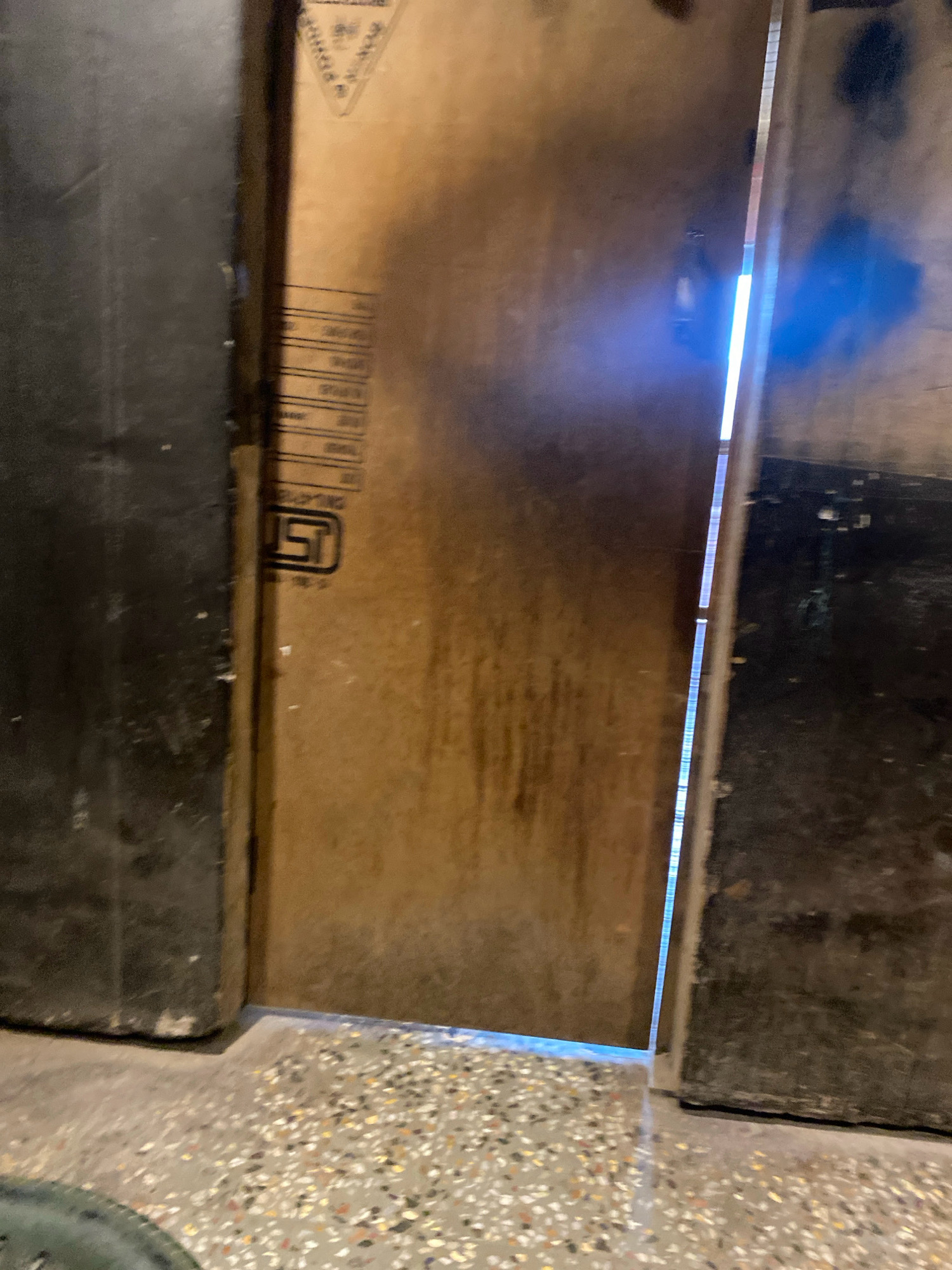 An interior scene within a building in Kodinar, India, features a narrow doorway between two large, dark, vertical objects that appear to be panels or doors. The object on the left is dark gray and textured, with visible scuffs and wear at its base. The object on the right is a lighter brown, possibly plywood, with printed text and a logo visible, including what appears to be an "ISI" mark. A bright blue light emanates from a thin gap between these two objects, creating a stark contrast. The floor in the foreground is a speckled, mosaic-like material. The lighting is dim, with the blue light being the primary light source.