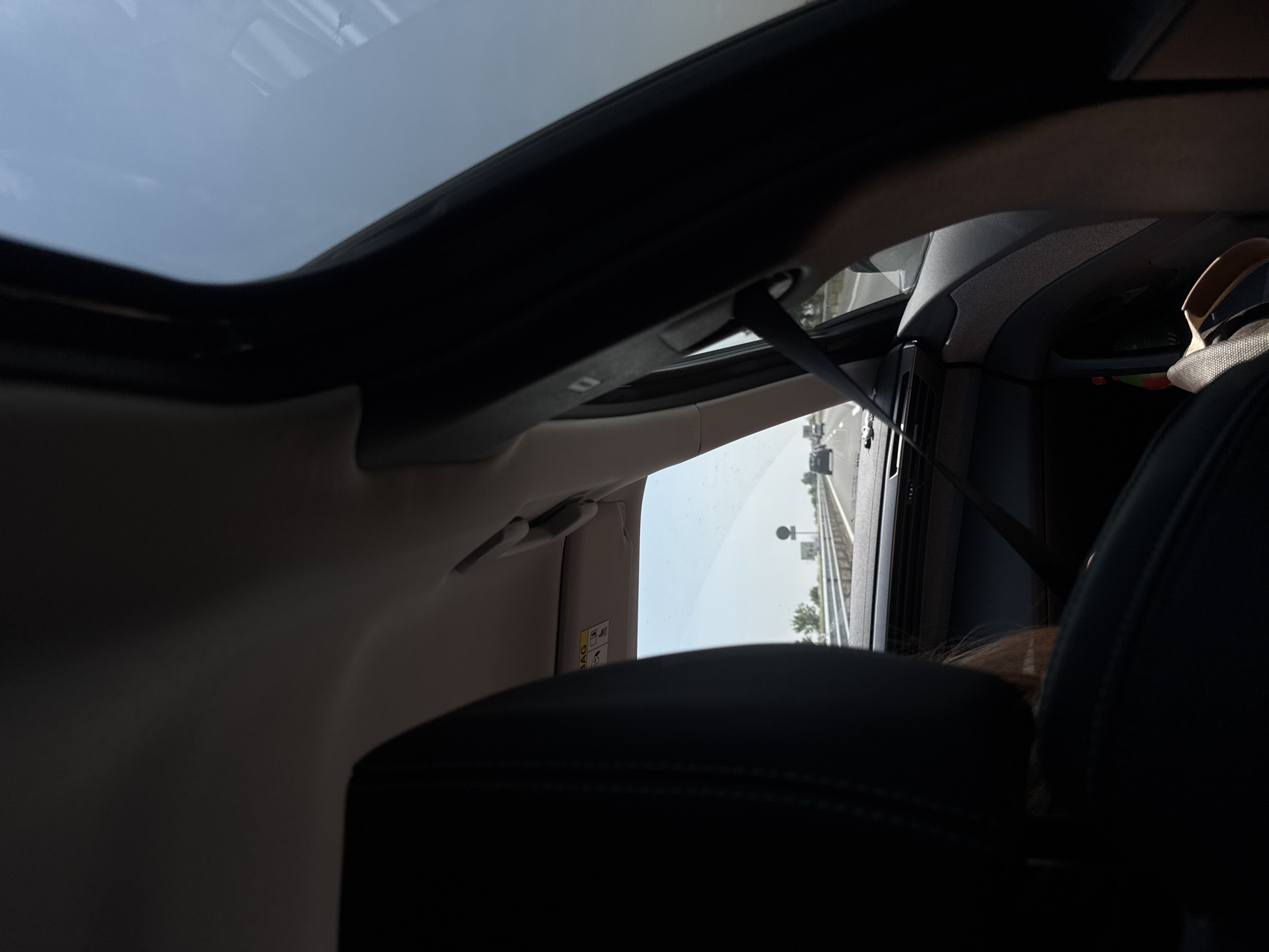 The image captures a forensic perspective from inside a moving vehicle, looking forward and slightly to the right, on a multi-lane highway near Malo Konare, Bulgaria, under a clear, bright daytime sky. The dark-colored rear of a front passenger seat dominates the left foreground, partially obscuring a person with light brown hair. To the right, the vehicle's interior trim, A-pillar, and a black seatbelt are visible. An upward-folded sun visor on the passenger side displays an "AIRBAG" warning label. On the floor or door pocket on the right, a light-colored sandal is present. Through the windshield, an asphalt highway stretches into the distance, populated by other vehicles. A metal guardrail lines the right side of the road, behind which a circular road sign and sparse vegetation are discernible.