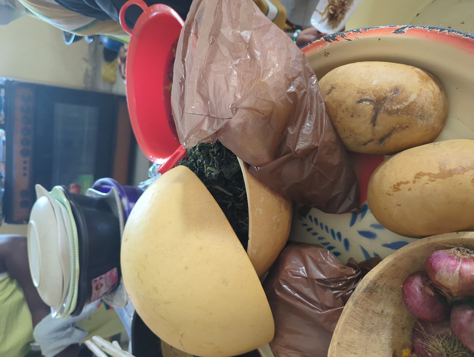 In an indoor setting in an unknown city, a view from above shows various cooking-related items. A cut calabash, or bottle gourd, sits near a red plastic bowl. A brown plastic bag is beside a wooden bowl of red onions. Other calabashes sit in a white bowl with blue accents. Additional bowls and cookware are visible in the background.