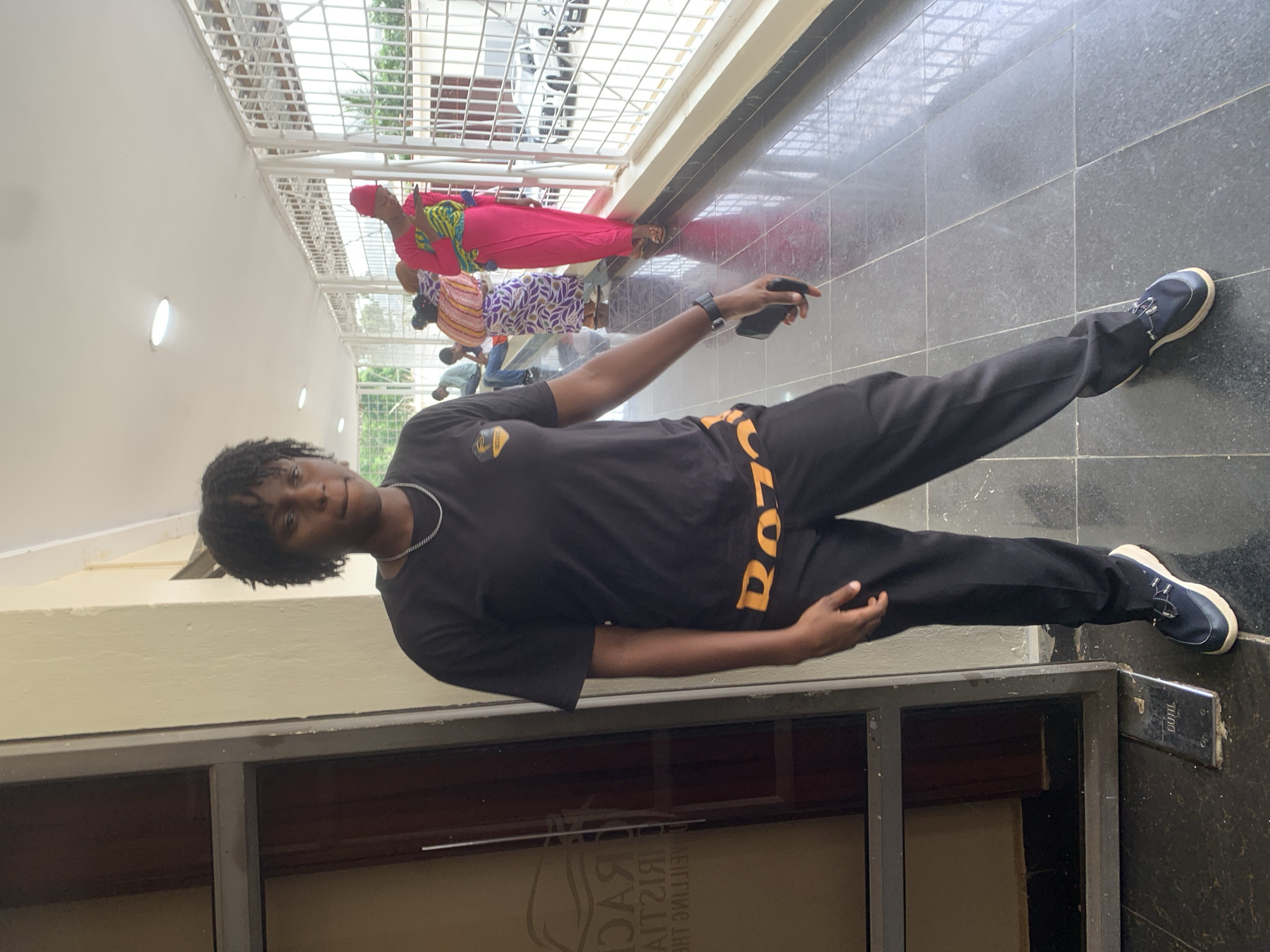 The image captures a young woman standing in a covered walkway, possibly in Abuja, Nigeria. She is wearing a black t-shirt with a yellow logo and black pants with yellow lettering. Her hair is styled in short dreadlocks, and she is wearing a silver chain necklace. She is holding a black mobile phone in her right hand.
In the background, several other people are visible, including a woman in a bright pink dress and headscarf, and another person in a patterned outfit. The walkway has a tiled floor and a white ceiling with light fixtures. A metal fence lines the right side of the walkway, offering a view of trees and buildings outside. To the left, a glass door with signage is partially visible. The overall lighting suggests it is daytime.