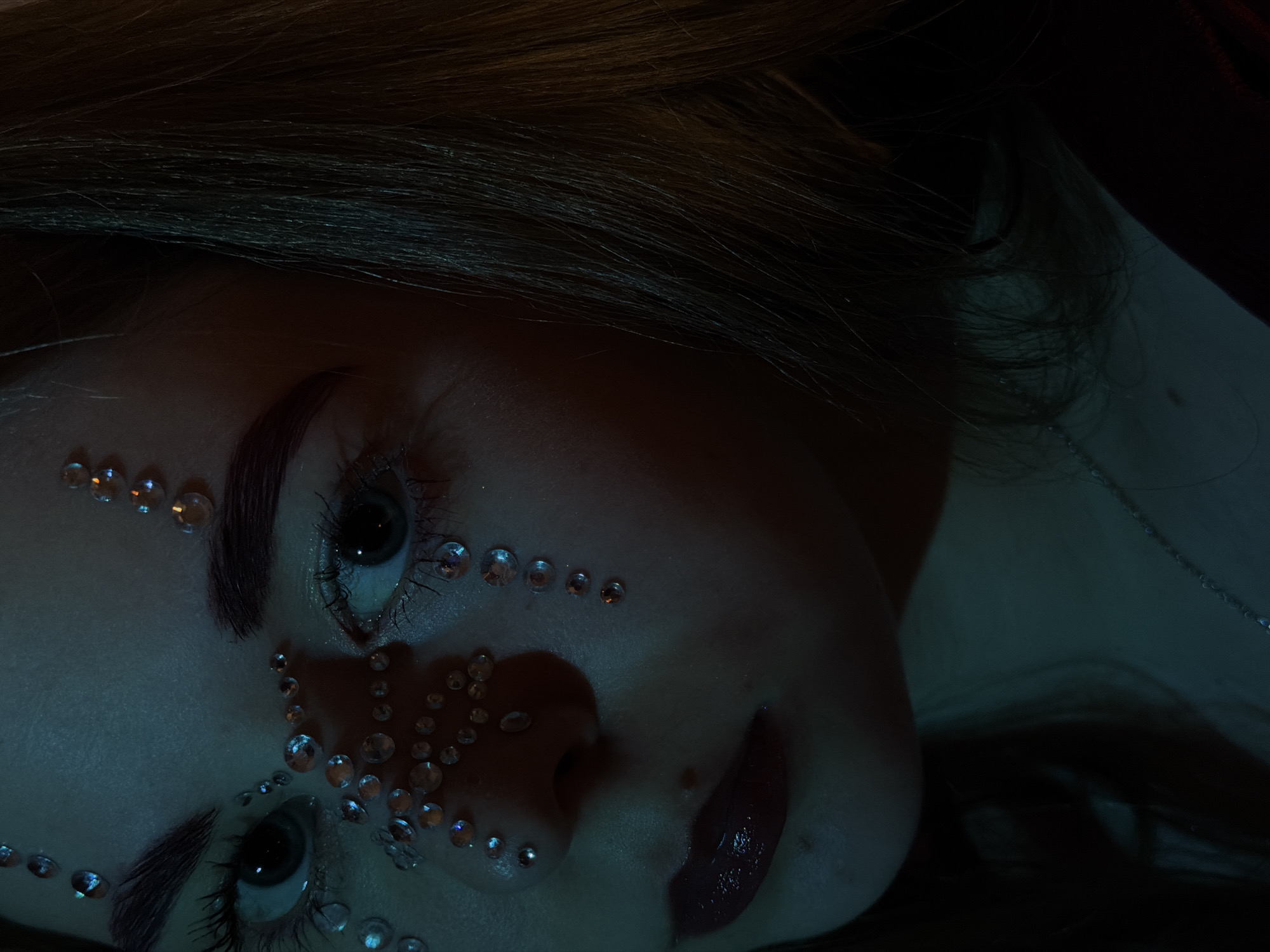 A close-up, eye-level shot depicts the face of a person with embellished facial makeup, featuring small, reflective gems adhered around the eyes, nose, and forehead. The subject has fair skin, dark eyebrows, and dark hair, with a glossy lipstick finish. The lighting is subdued, with a cool, blueish tone dominating the color palette. A hint of what appears to be a collarbone and a thin necklace can be seen on the side. Without further information, the location in Perm, Russia, cannot be determined from the image.