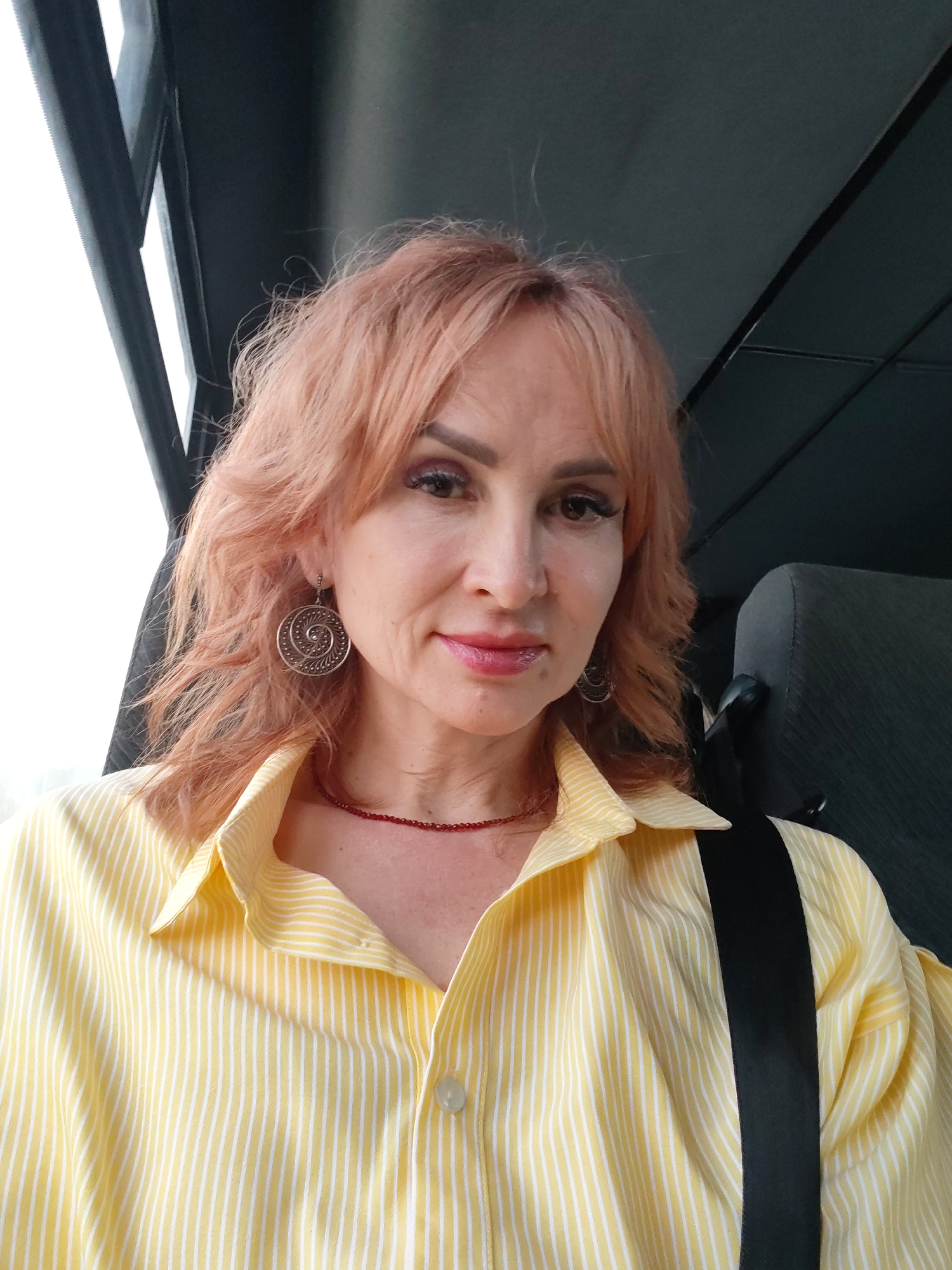 A medium eye-level shot depicts a light-skinned woman with reddish-blonde, shoulder-length hair and bangs. She is seated inside a vehicle, wearing a yellow and white striped button-down shirt and a red beaded necklace. A black seatbelt crosses her upper body. She wears ornate, circular earrings. The backdrop is comprised of a vehicle interior, with a window on the left and a headrest in the top right corner. The location is Chaikovsky, Russia, based on image context.
