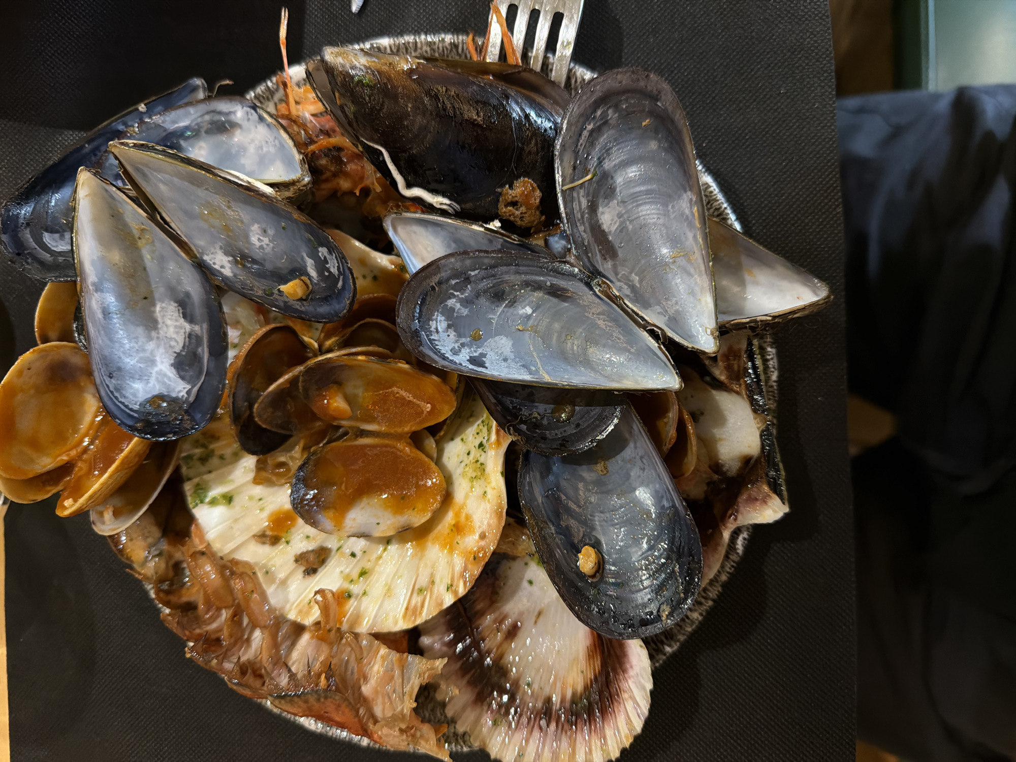 The image shows a plate of seafood, likely served as tapas in Santiago De Compostela, Spain, given the regional cuisine. Dominating the view are numerous empty mussel shells with their characteristic blue-gray interior. Alongside the mussels are open scallop shells with remnants of cooked seafood. Also visible are several clams, bathed in a brown sauce. A metal fork is partially visible in the plate. The plate sits on a dark surface, possibly a restaurant table.