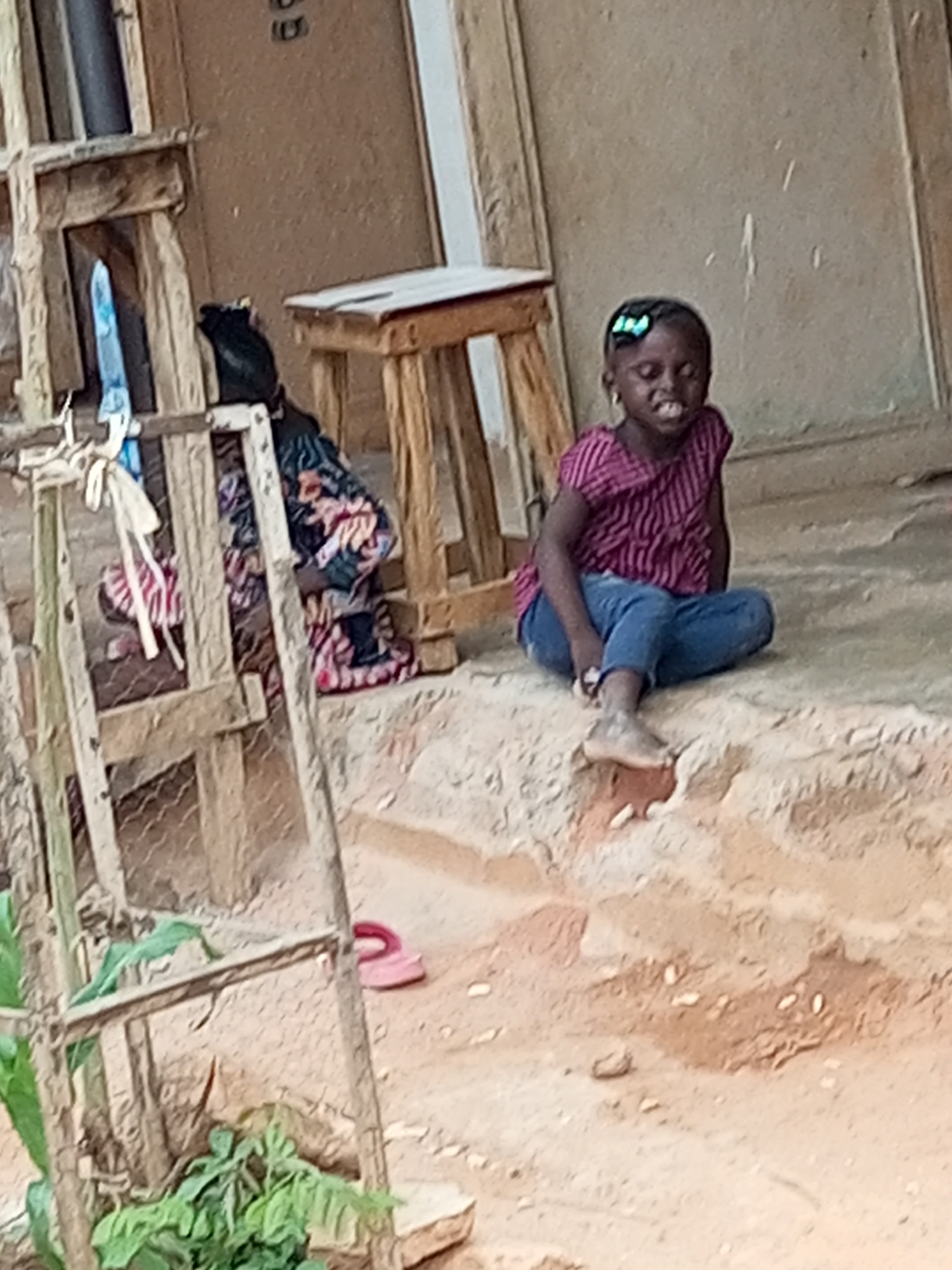 A young Black girl with a blue bow in her hair sits cross-legged on a dirt embankment. She is wearing a magenta and black striped t-shirt and blue jeans. Her mouth is open in a wide smile, and her eyes are closed. Behind her, a wooden structure appears to be some sort of table or bench. To the left, a person is partially visible, wearing a patterned dress and sitting on the ground. The environment is a dusty, outdoor area with a building in the background. The overall scene suggests a casual, everyday moment in a residential or public space in Tempure, Nigeria.