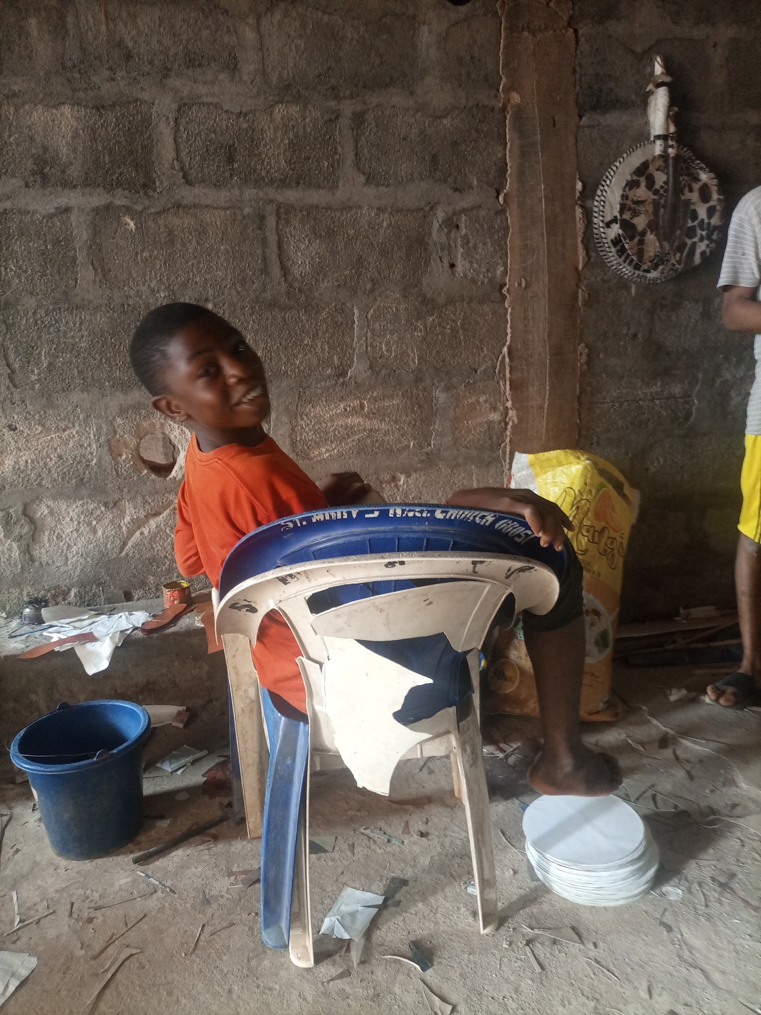 A young boy wearing an orange shirt is sitting in a blue and white plastic chair, facing the camera. He is smiling and looking to his left. Behind him, a rough brick wall is visible. To the left of the boy, a blue bucket is on the floor. To the right of the boy, a large yellow bag is leaning against the wall, and a stack of white circular objects is on the floor. Another person's legs and feet are visible on the right side of the image. The scene appears to be indoors, in a dimly lit space with a dirt floor.