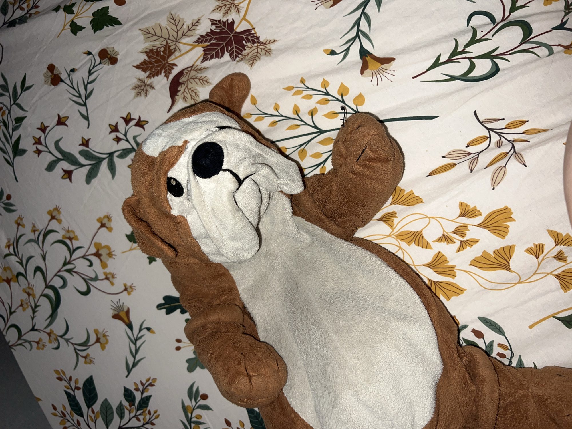 A brown and white plush dog toy lies on a patterned bedsheet. The bedsheet has a white background with a repeating pattern of green and brown foliage. The dog toy is positioned on its back. No people or other objects are visible. The location in Chaikovsky, Russia is not determinable from this image.