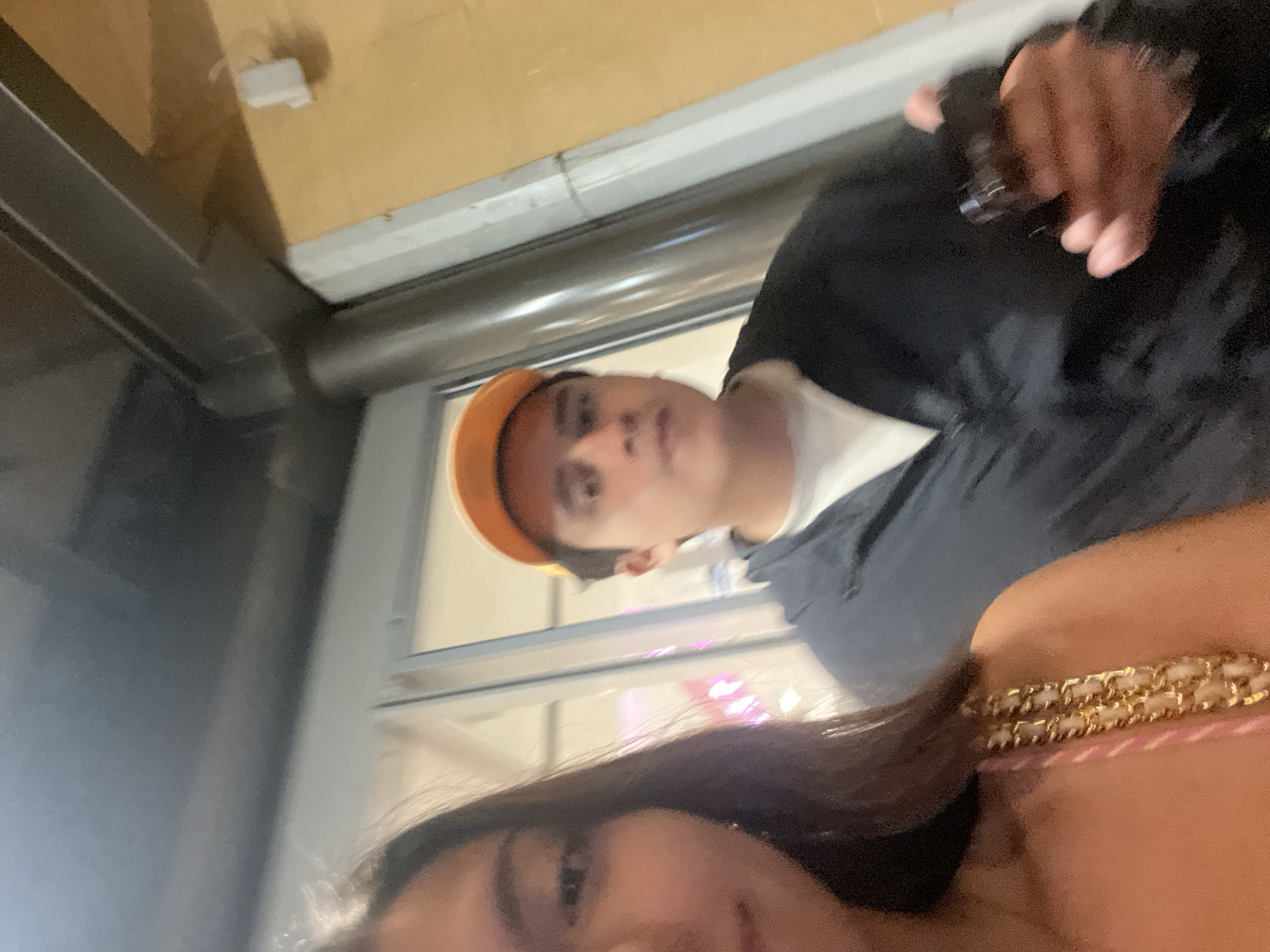 A selfie taken indoors with two people. A person with brown hair and light skin is partially visible in the foreground wearing a pink top and a gold chain necklace. Behind them, a person is wearing an orange hat, a white t-shirt, and a black jacket, appearing to hold a dark object. The background shows a window with metallic frames, outside of which indistinct lights and a building facade can be seen. A piece of the ceiling and wall is visible in the upper left of the frame, which are tan in color, along with a small rectangular white object. Location is suspected to be Puebla, Mexico.