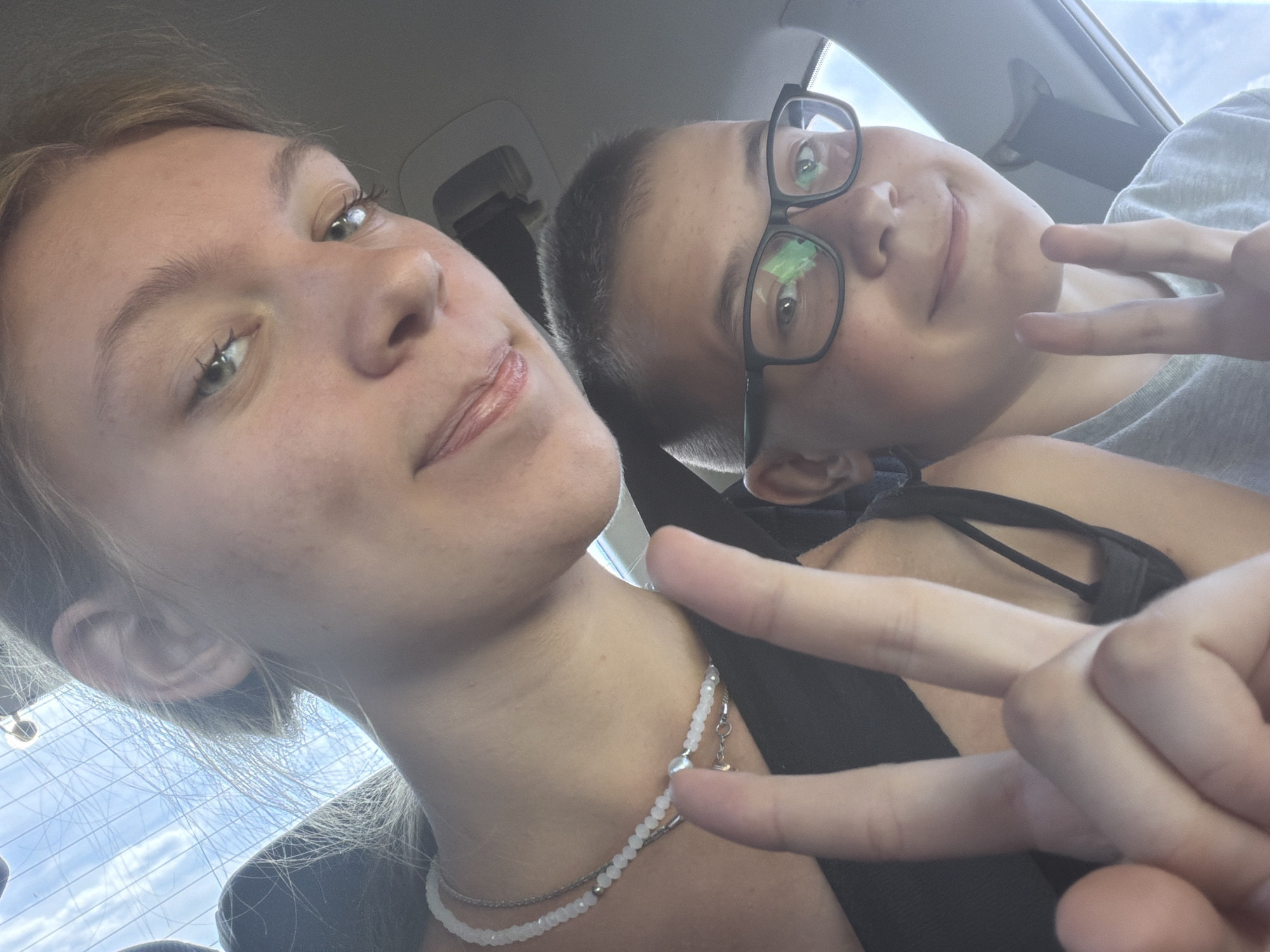 In the city of Novoe Feizullovo, Russia, two individuals, a young woman and a boy, are captured in a selfie taken from inside a vehicle. The young woman is in the foreground, with her face partially visible as she looks towards the camera. She has light-colored hair, and is wearing a necklace. Her hand is raised, displaying a peace sign. Beside her, and slightly further back, is a boy wearing glasses. He also has his hand raised, forming a peace sign. The background suggests the interior of a car, with a window and the sky visible. The overall impression is that of two individuals enjoying a moment together, likely during travel.