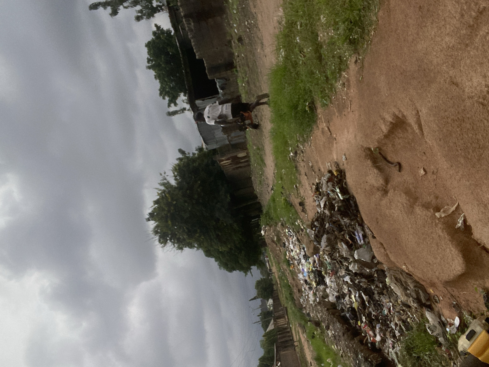 The media file depicts an outdoor scene in Gbaza, Nigeria, characterized by a dirt road, overgrown grass, and a pile of garbage. The image is oriented vertically, with the ground and sky appearing on the left and right sides, respectively.
In the center of the frame, a person wearing a white shirt and dark shorts is walking along the dirt path. The individual appears to be carrying something orange in their arms. Behind the person, there are some simple structures, possibly homes, made of corrugated metal and brick. Lush green trees are visible in the background, contrasting with the arid ground.
To the right of the dirt road, a significant accumulation of trash is visible, consisting of various plastic containers, bags, and other discarded items. This suggests a poorly managed waste disposal area. A small, yellow object, possibly a toy or a piece of equipment, is partially visible in the lower right corner of the image, near the garbage pile.
The sky is overcast with heavy grey clouds, indicating a possibility of rain or generally gloomy weather. The lighting is diffused, suggesting it is either early morning or late afternoon, or the overcast conditions are the primary factor. The overall impression is one of a somewhat neglected urban or semi-urban environment, where daily life continues amidst environmental challenges. No visible text is present in the image.