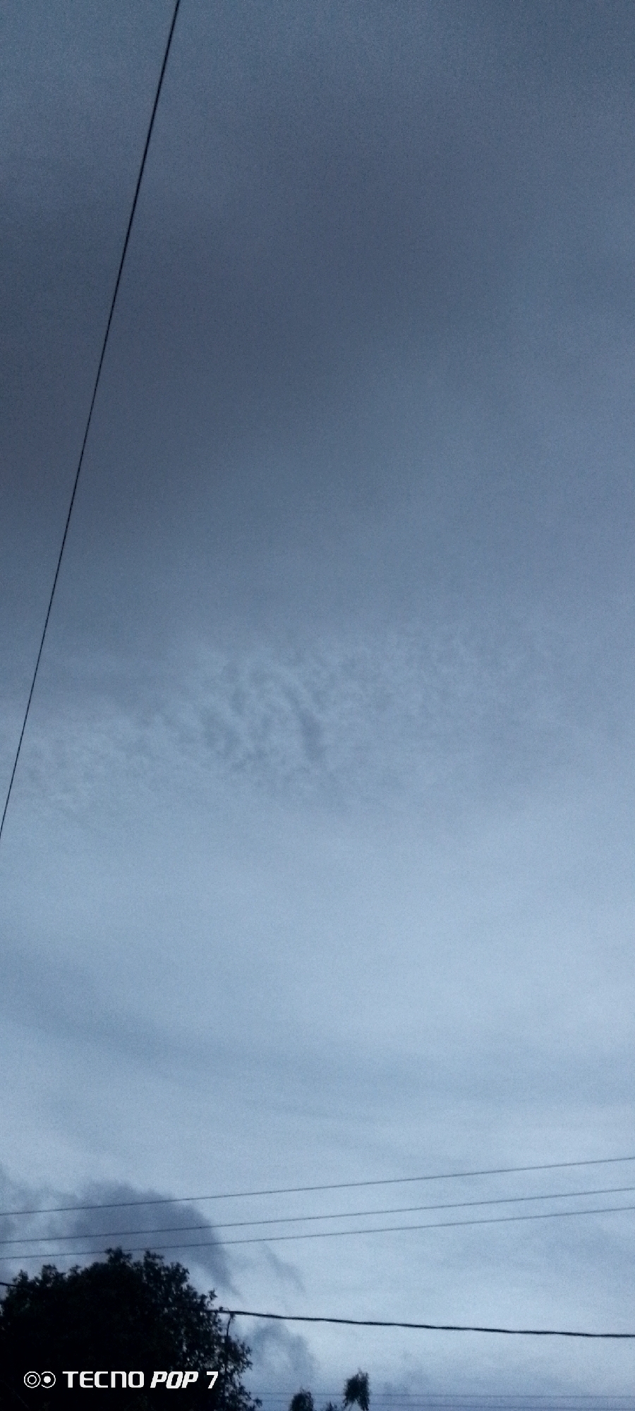 The image shows a cloudy sky with a few trees and power lines visible at the bottom. A single, dark cable runs diagonally from the upper left corner towards the center. The lower portion of the sky is slightly brighter. The bottom of the image also shows the text "TECNOS POP 7". Based on the model mentioned in the image I am assuming this was taken from Altagracia, Venezuela.