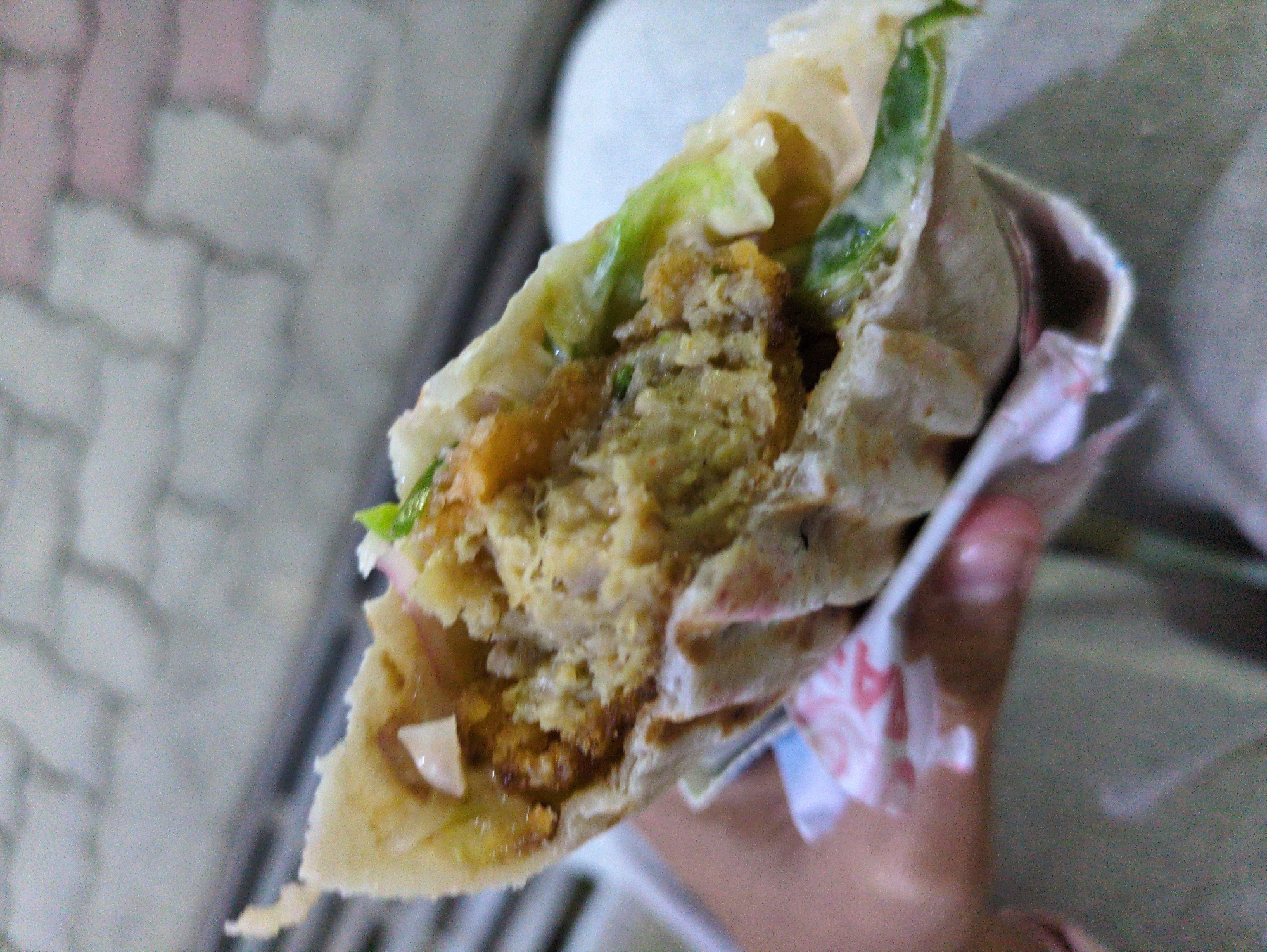A close-up, slightly blurred shot shows a person holding a falafel wrap. The wrap is made with a thin flatbread, filled with falafel patties, lettuce, and what appears to be a white sauce and red onion. The background is out of focus, showing a cobblestone-like surface, possibly a pathway. The scene is outdoors, likely during daylight hours. The image does not provide specific information about the location within Dharamshala, India.