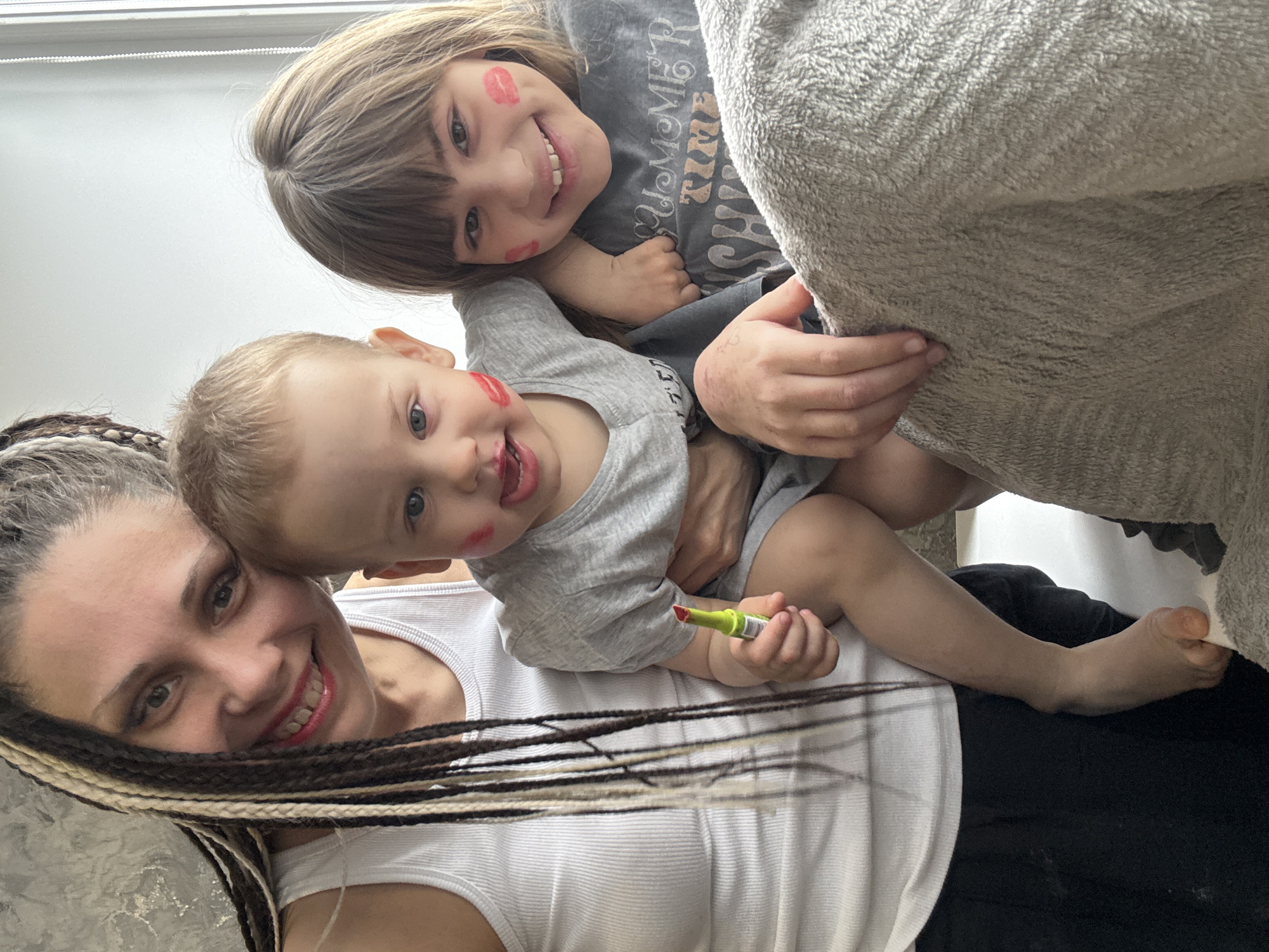 The image captures a joyful indoor family moment, likely a selfie, featuring an adult woman, an older child, and a baby/toddler. The photo is taken from a top-down perspective, with the subjects appearing to be lying down or arranged closely together.
The adult woman, positioned at the bottom left, has long, dark hair styled with lighter, possibly blonde, braids. She is wearing a white ribbed tank top and smiles warmly at the camera, revealing red lipstick on her lips. She is holding the baby.
Above her, an older child, likely a girl, smiles broadly at the camera. She has shoulder-length light brown hair with bangs and is wearing a grey t-shirt that clearly displays the text "SUMMER TIME" in stylized lettering. Several red lipstick kiss marks are playfully smeared across her right cheek. She is nestled close to the woman and a large, grey, fuzzy blanket or cushion that occupies part of the upper right of the frame.
To the right of the woman and below the older child, a baby or toddler looks towards the camera with a wide, happy smile. The baby has light hair and is also wearing a grey t-shirt. Similar to the older child, the baby has red lipstick marks on his left cheek and around his mouth, suggesting they have been playing with makeup. He holds a bright green lipstick tube with the red lipstick extended.
The setting appears to be a bright room, possibly with a window providing ample natural light, visible as a bright white area in the top left background. The overall mood conveyed by the smiling faces and playful lipstick application is one of happiness and family fun in a domestic environment in Rusanovo, Russia.