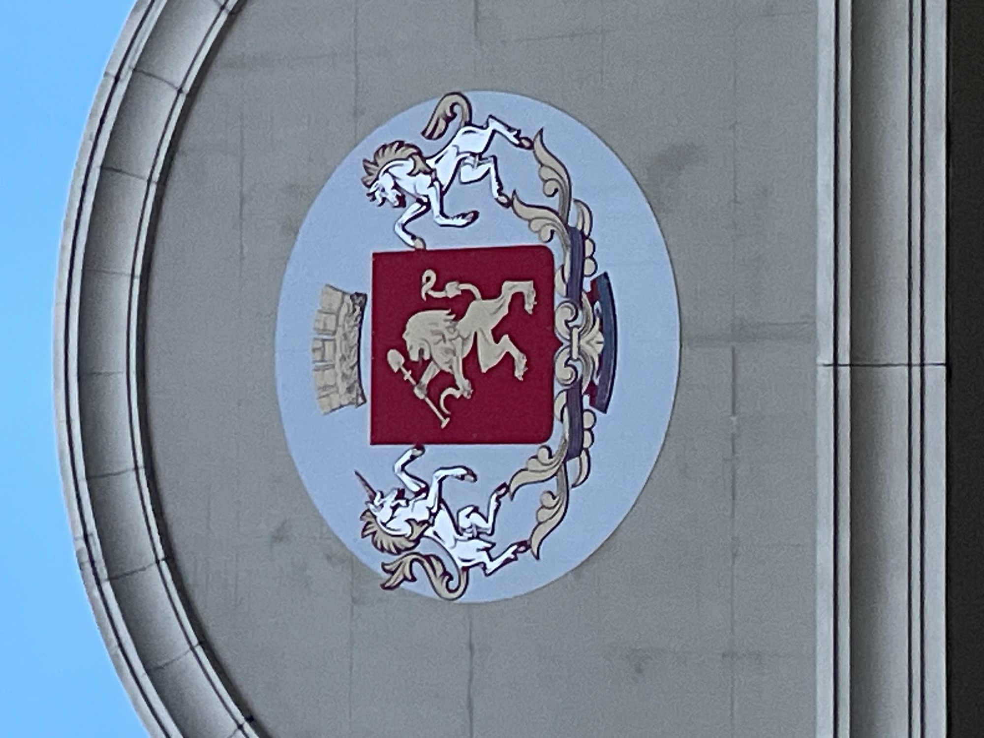 The image shows a circular coat of arms affixed to a building. The coat of arms features a red shield on which a golden lion rampant holds a golden shovel and sickle. To the left of the shield is a golden tower. The coat of arms is surrounded by two white unicorns rampant and ornamented scrollwork. The coat of arms is located on a gray building in Krasnoyarsk, Russia.