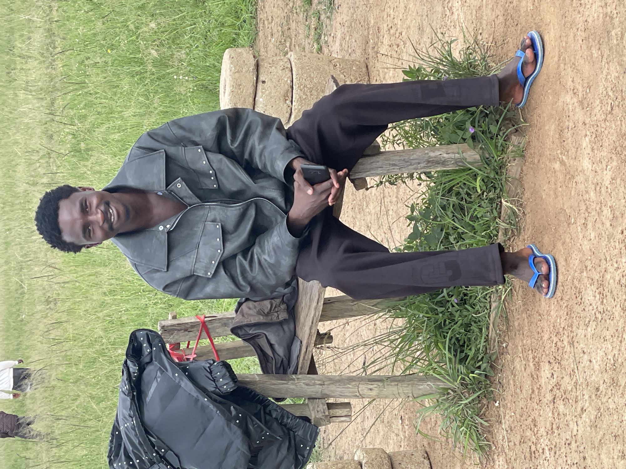 A Black man sits on a rustic wooden bench in Durumi, Nigeria. He is wearing a grey leather jacket over a dark shirt, black pants, and blue flip-flops. He is holding a black mobile phone in his hands, which are clasped together. The bench is positioned outdoors on a dirt path, with tall green grass and vegetation behind it. To the left of the bench is a dark, puffy jacket draped over a wooden support. To the right of the bench are three stacked, round, light-colored stones. In the background, two other individuals are visible walking in a grassy field.