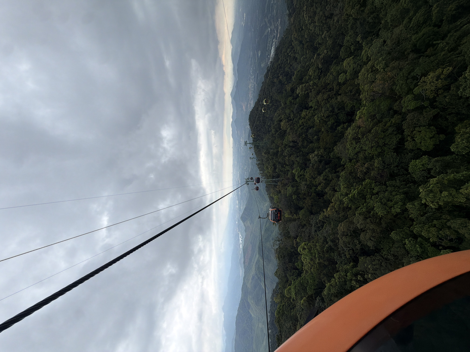 A high-angle, slightly distorted photograph captures a cable car ascending over a lush, dense forest in Đà Nẵng, Vietnam. The cloudy, overcast sky fills the upper left quadrant of the image. In the center, a thick, dark cable stretches upwards, supporting several gondola cars. A few are visible in the distance, and one, closer, has a predominantly red or orange exterior. The forest extends from the right edge toward the center of the image. Mountains stretch from above the forest on the right to the center horizon, fading into a hazy or smoggy view of the distant land. In the lower right corner, a section of an orange or brownish structure is visible.