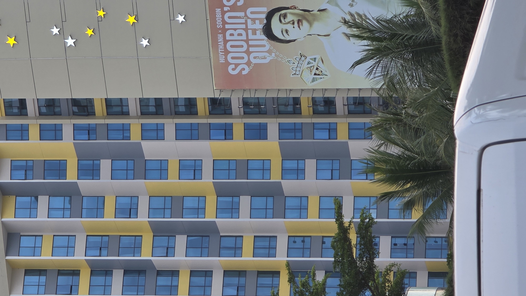 This image captures a vibrant, modern urban scene, likely in Nha Trang, Vietnam, on a bright, sunny day. The central focus is a multi-story building with contemporary architecture, featuring a striking facade of alternating rectangular sections in yellow, grey, and white/light grey. Numerous rectangular windows with blue-tinted glass are evenly spaced across the building's surface.
On the upper section of the building, a large vertical billboard displays a portrait of a person, likely an idol or celebrity. The individual has light skin and dark hair, is wearing a white top, and sports a prominent, ornate diamond-shaped pendant around their neck. Visible text on the billboard reads "HUY THANH x SOOBIN" and "SOOBIN'S QUEEN," suggesting a promotional collaboration or fandom reference.
Above the building and billboard, an overhead structure is adorned with decorative star-shaped lights, alternating between yellow and white. To the right, lush green palm trees and other foliage partially obscure the building, adding a tropical touch to the urban setting. The far right edge of the frame shows a portion of a white vehicle, possibly a van or bus, indicating a busy street or thoroughfare. The clear lighting suggests it is daytime.