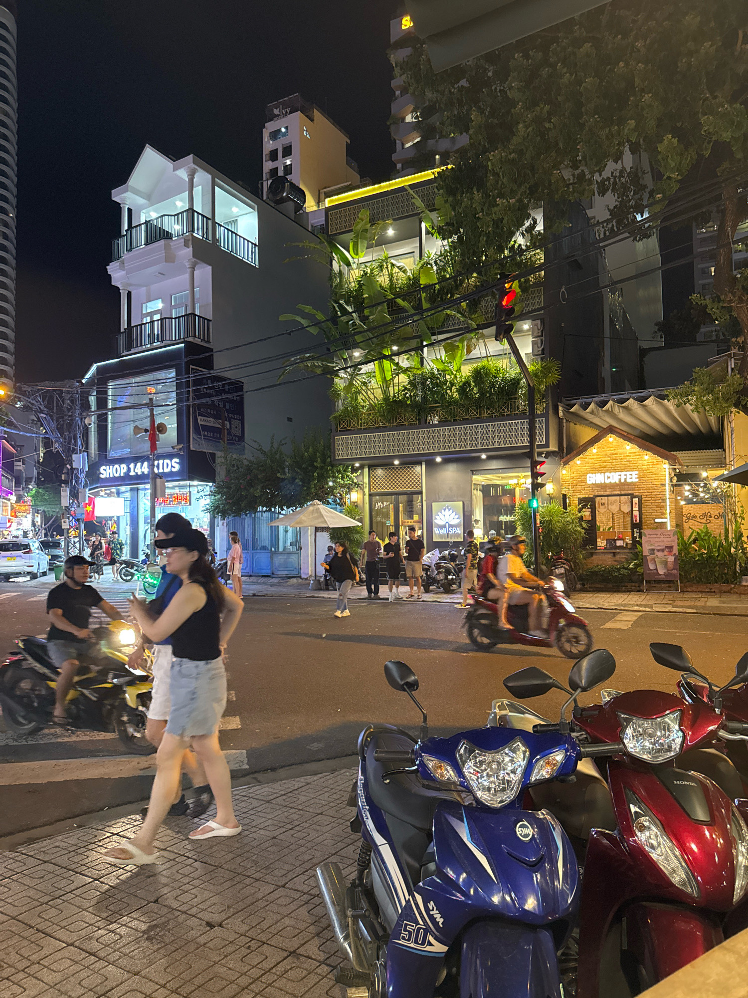 This is a nighttime street scene in Nha Trang, Vietnam, showing a busy urban environment with shops, cafes, and traffic. In the foreground, several motorcycles are parked along the sidewalk. A young couple walks on the sidewalk, with the woman in a black tank top and denim shorts, and the man in a black t-shirt and shorts, both wearing sandals. They are in the process of crossing a street. A yellow motorcycle with two riders is in motion behind them. Across the street, a brick building with a pitched roof and "GIN COFFEE" signage is illuminated. Next to it, a modern building with multiple levels features a facade of lush greenery. Further down the street, a shop labeled "SHOP 144 KIDS" is visible, along with other businesses and apartment buildings. Traffic lights are visible, indicating ongoing vehicle and pedestrian activity. The overall atmosphere is lively and characteristic of a bustling Asian city at night.