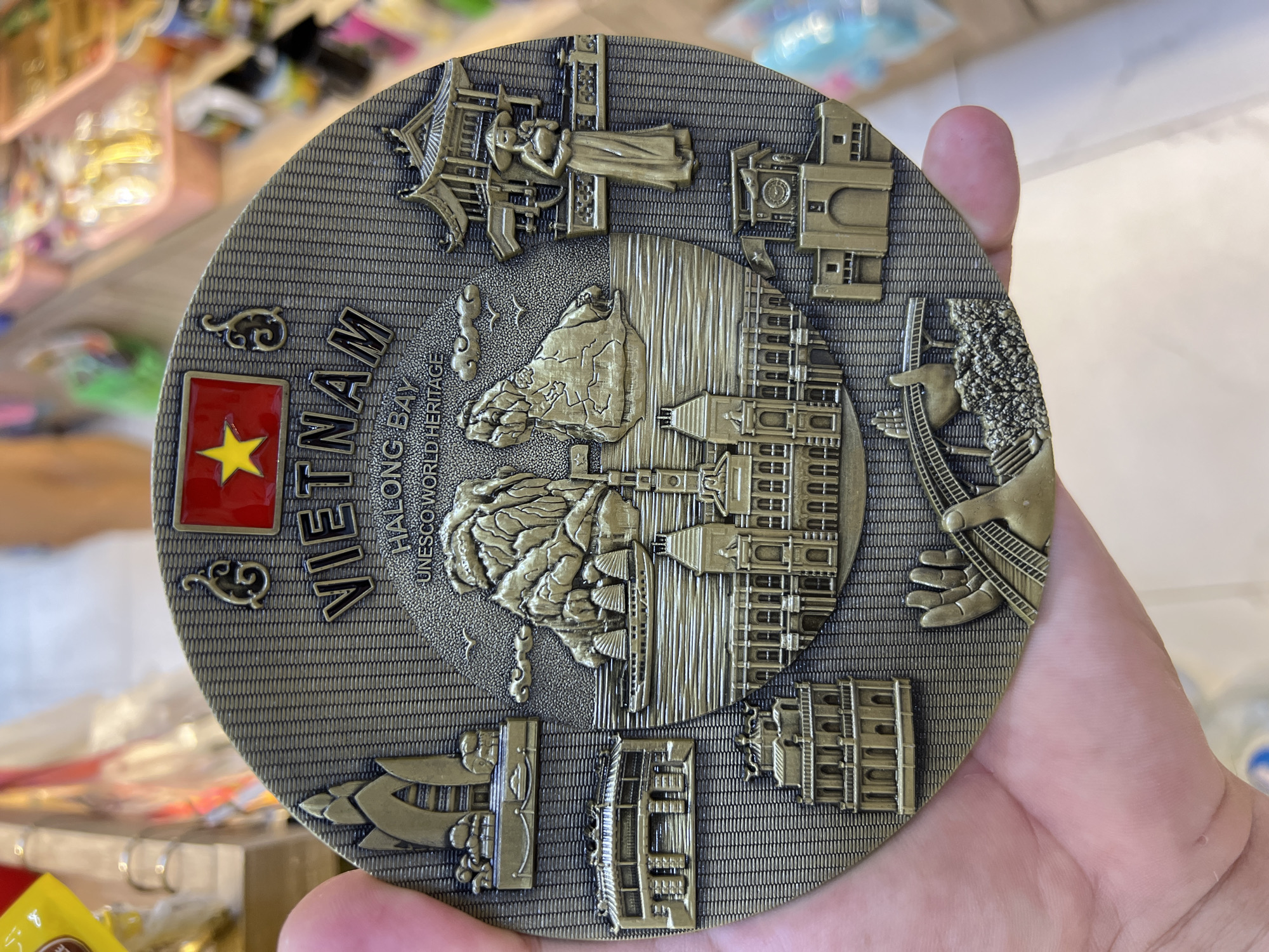 A circular souvenir magnet for Vietnam is held in a person's hand. The magnet features embossed relief images of Vietnamese landmarks and a woman in traditional attire. The words "VIETNAM," "HALONG BAY," and "UNESCO WORLD HERITAGE" are visible. The background of the image is blurred, showing shelves with various items, suggesting a shop environment. The magnet itself has a metallic, antique bronze finish.