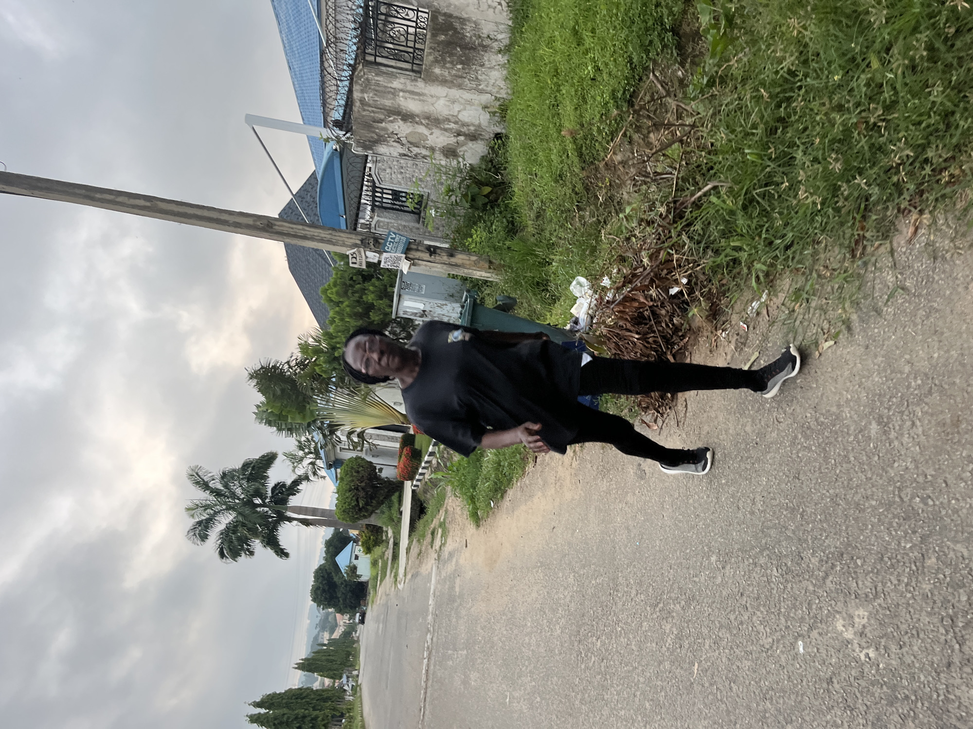 The image depicts a woman running on a paved road in Abuja, Nigeria. She is wearing a black t-shirt, black leggings, and black and white running shoes. The road appears to be in a residential area, with houses and trees visible in the background. A prominent utility pole stands on the right side of the road, and there is a drainage ditch with overgrown vegetation along the side. The sky is overcast with some patches of light. The overall scene suggests an outdoor activity in a suburban environment.