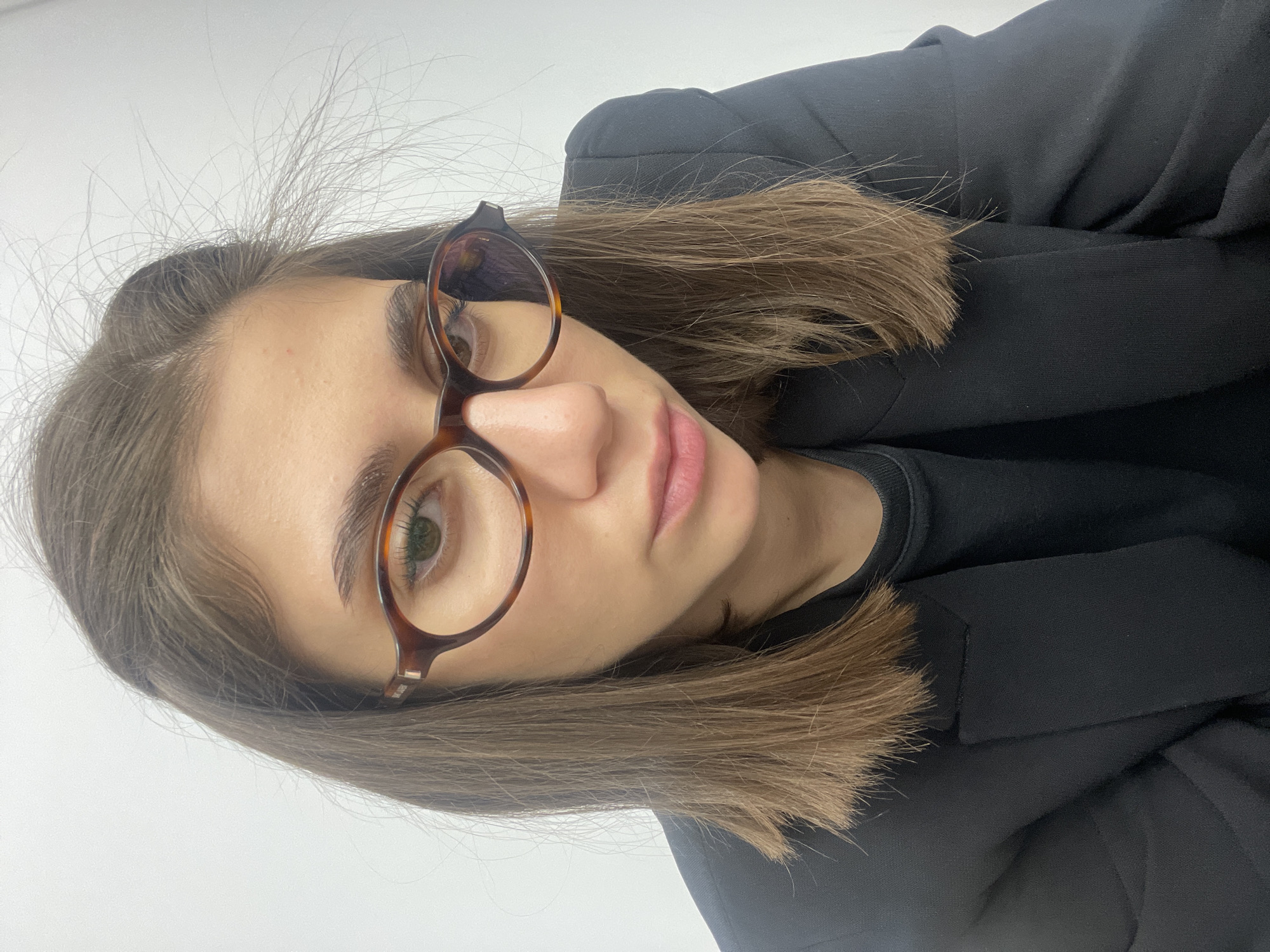 A close-up, rotated portrait features a young woman with medium brown, shoulder-length hair and green eyes, wearing roundish tortoiseshell-framed glasses. Her head is tilted, and she looks directly towards the camera with a neutral expression. Her hair appears slightly disheveled with some flyaways visible around the crown. She is dressed in a black jacket or blazer over a black top, with the collar of the inner garment just visible. The background is a plain, light-colored surface, likely a white or light grey wall, indicating an indoor setting. The lighting is bright and even. There are no specific activities depicted beyond posing for the photograph. No text is visible in the image. The location context is Sofia, Bulgaria.