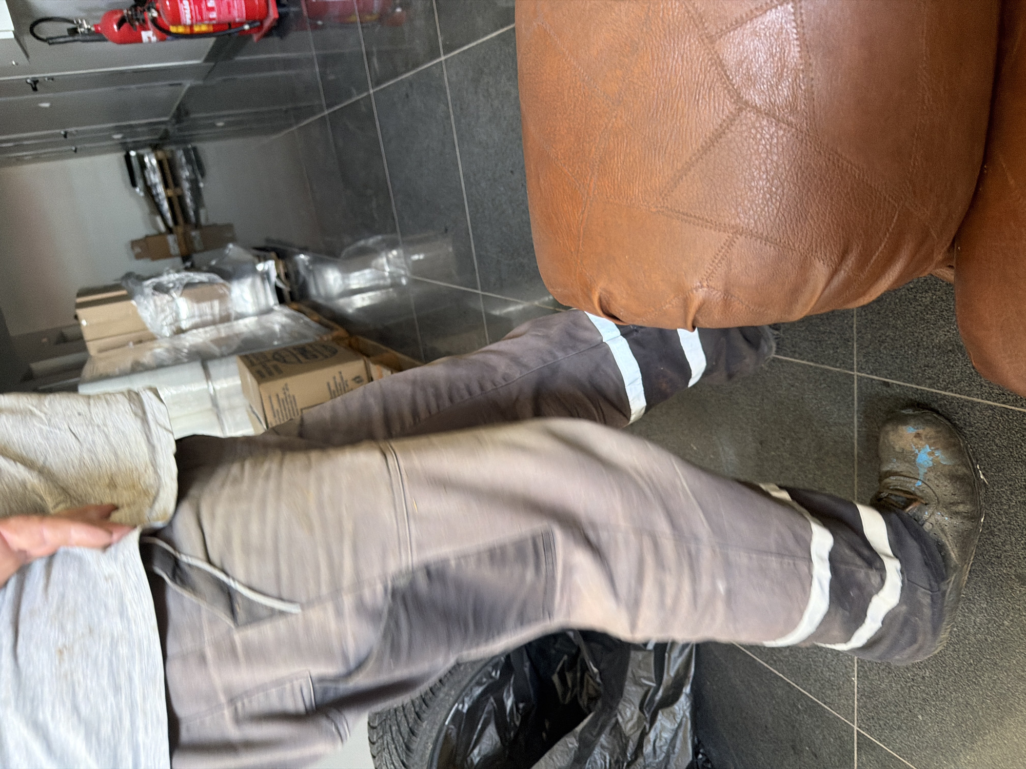 The image shows a low-angle shot of a person's lower body and legs. The person is wearing work pants with reflective white stripes around the lower calves and muddy work boots. A portion of a brown leather cushioned seat or ottoman is visible in the foreground, partially obscuring the person's right leg and foot. In the background, there is a hallway with a tiled floor, doors, and stacked boxes. To the right, a red fire extinguisher is mounted on a wall. The scene appears to be an interior space, possibly a storage area or corridor, within a building. There is no visible indication that this location is in Dolno Ablanovo, Bulgaria.