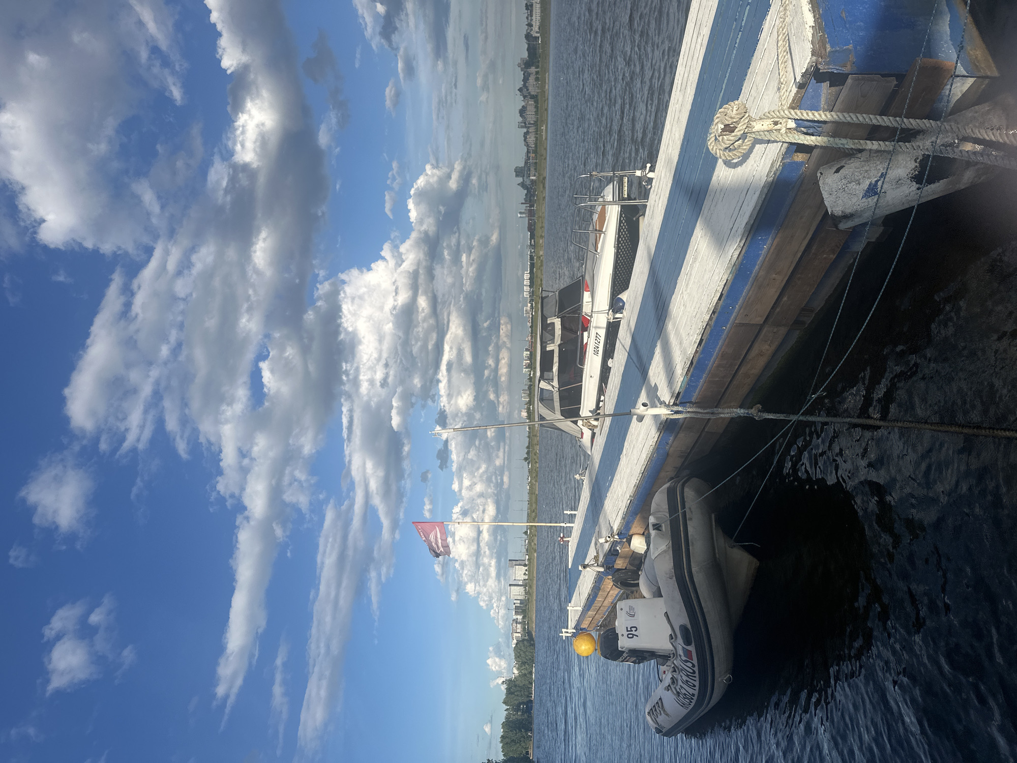 The image shows a boat docked on a body of water, likely the Volga River, in Kazan, Russia. The boat features blue and white striping. A smaller inflatable dinghy is attached to the side of the boat. The dinghy has the number "95" visible and a Russian flag sticker. There is a Russian flag flying from a pole on the boat. In the background, a cityscape is visible with various buildings. The sky is blue with scattered white clouds. The water appears calm.