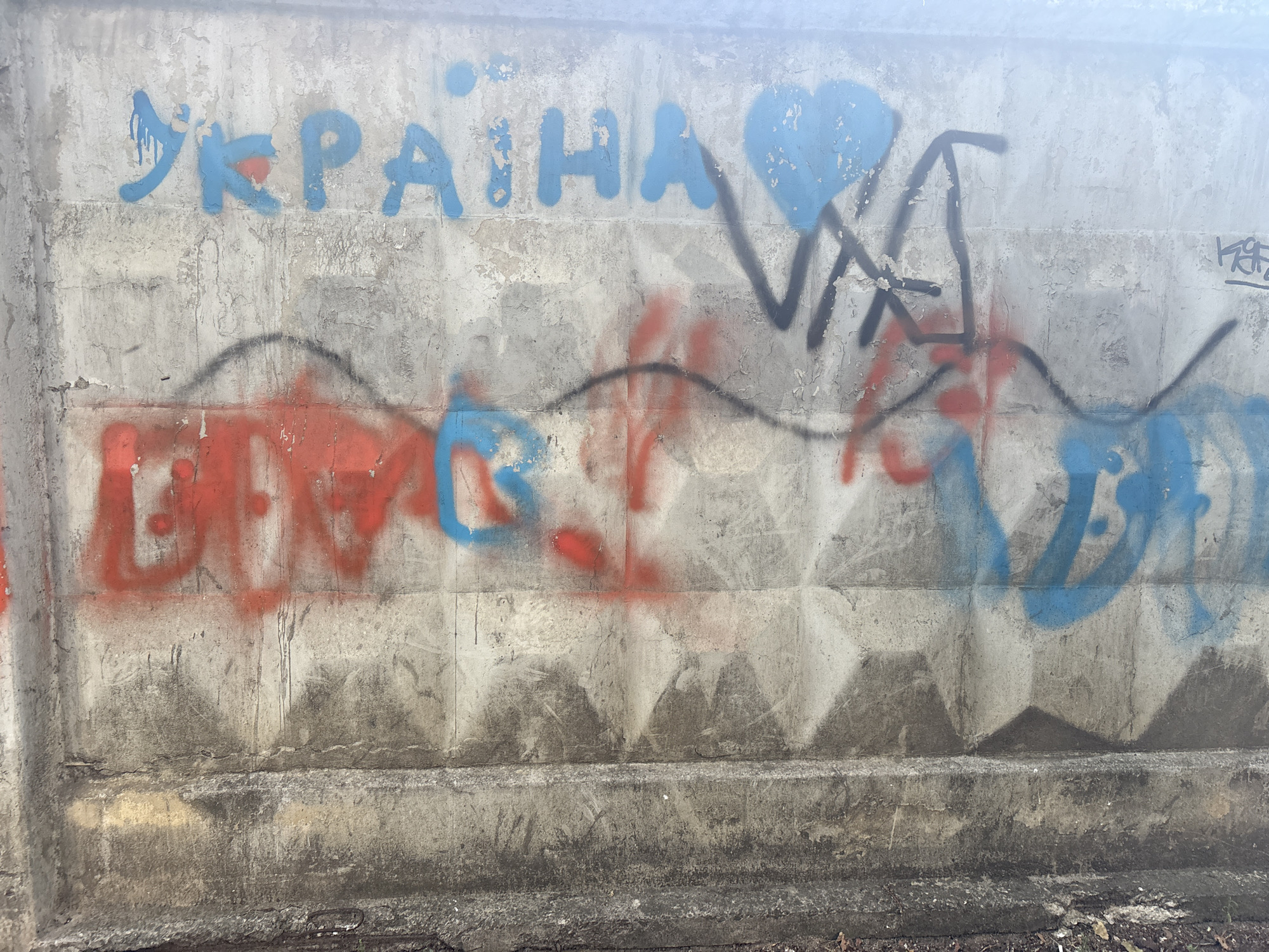 The image depicts a concrete wall covered in graffiti. The graffiti is primarily in blue and red spray paint. The word "УКРАЇНА" (Ukraine) is written in large blue letters across the top portion of the wall. To the right of the word, there is a blue heart shape. Below this, red and blue spray paint forms abstract shapes and lines, appearing as wavy patterns and possibly lettering that is difficult to decipher. The concrete wall itself is textured and appears weathered, with visible vertical seams or panels. The lower part of the wall shows signs of dirt and grime. The environment appears to be outdoors, with the wall likely part of an exterior structure. The image has a slightly hazy quality, possibly due to lighting conditions or atmospheric effects.