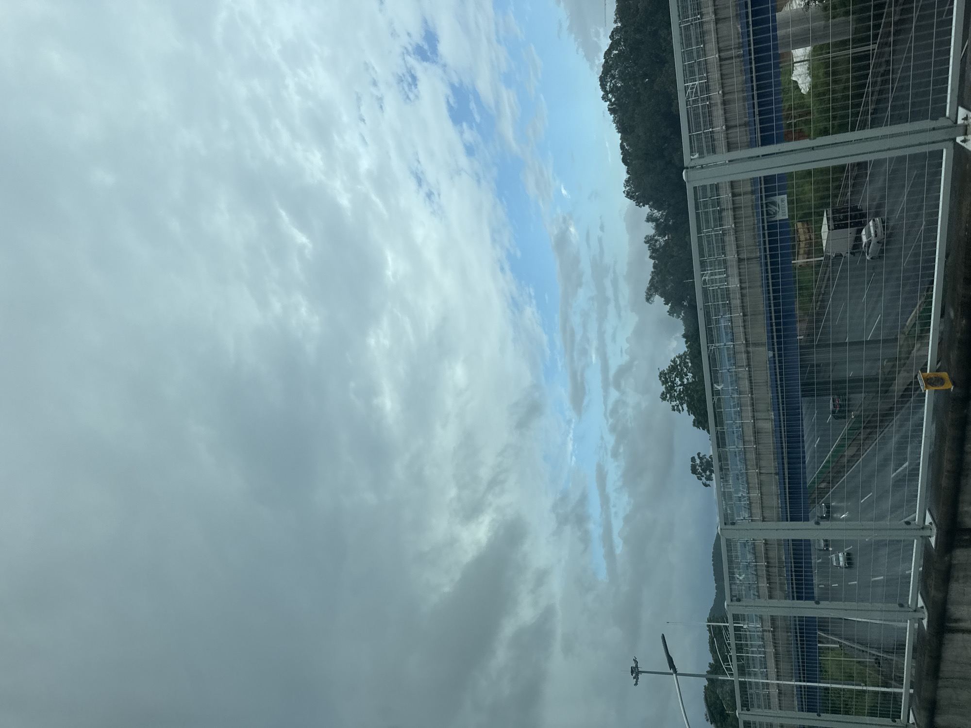The image captures a view of a multi-level highway interchange, likely in Longgang District, China, under a cloudy sky with patches of blue. The foreground features a wire mesh fence. Below the fence, a highway with multiple lanes is visible, with several cars and a truck driving on it. The road appears wet, suggesting recent rainfall. In the mid-ground, another level of the highway structure is evident, supported by concrete columns. Beyond the interchange, a dense line of trees forms a backdrop, with hills visible further in the distance. A tall light pole stands on the left side of the image. The overall atmosphere is overcast, with the clouds dominating the upper portion of the frame.