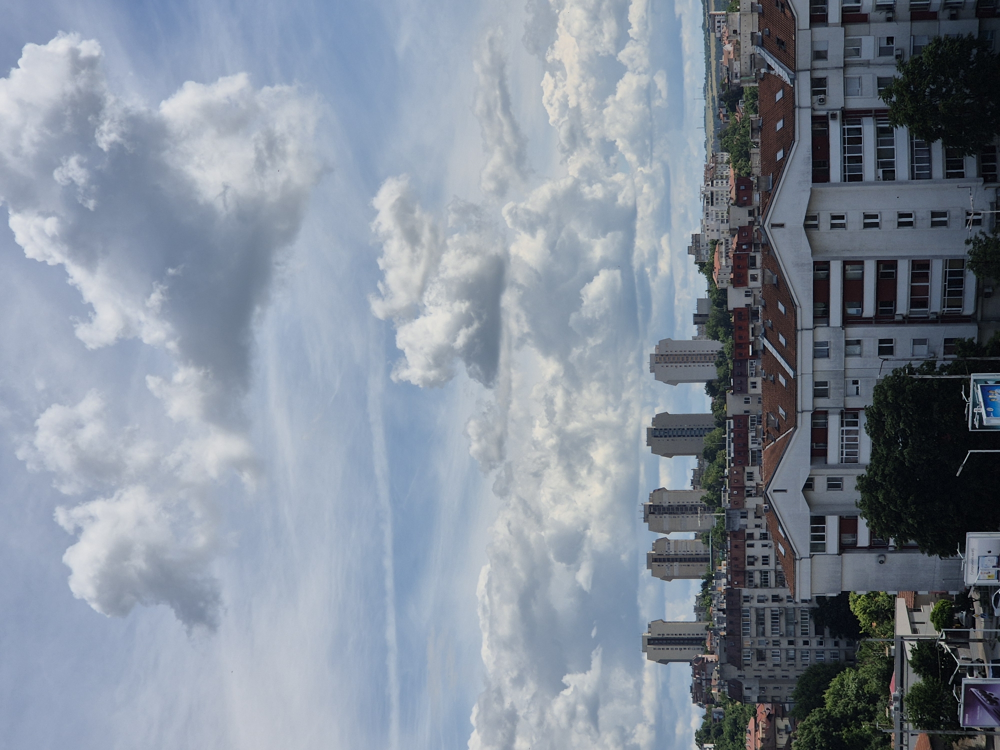 An aerial shot captures a densely built urban landscape under a sky filled with dramatic clouds. In the foreground, a large, multi-story apartment building with a white facade and reddish-brown tiled roof dominates the view. This building features numerous windows and balconies, and it extends across much of the frame. Behind it, a network of smaller buildings, also with tiled roofs, forms a dense urban fabric. Further in the background, a row of taller, distinct apartment blocks with pale facades and dark windows stand against the horizon, appearing as a more modern architectural element in the city. The sky is a dynamic mix of bright blue patches and large, billowing white and grey clouds, suggesting an overcast or partly cloudy day. Some greenery is visible, particularly trees interspersed among the buildings, especially on the left side of the image and near the foreground building. The overall scene depicts a residential area within a city, characterized by varying architectural styles and a prominent sky.
