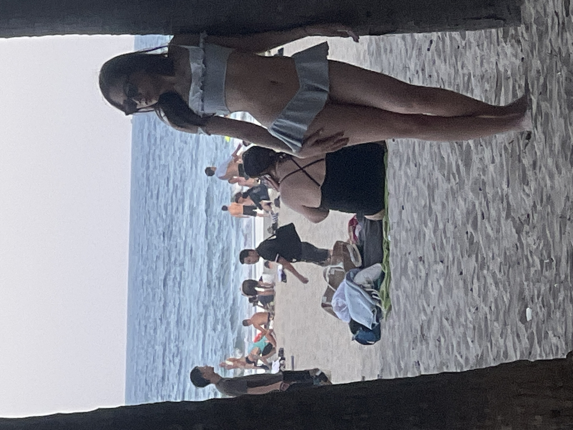 The image shows a group of people on a sandy beach next to the ocean. The perspective is from underneath a structure, looking up and out at the scene. A woman in a light-colored bikini is prominently featured in the foreground, lying on the sand. She is wearing sunglasses and her hair is dark.
In the background, several other individuals are visible, some sitting and others standing. The ocean is a calm expanse of blue water, meeting the shore under an overcast sky, suggesting it might be early morning or late afternoon. The sand is light-colored and appears slightly damp. The overall atmosphere appears relaxed, typical of a beach day.
There is no visible text in the image. The location cues are limited to the general beach setting, but the request specifies Đà Nẵng, Vietnam, which aligns with a coastal environment. The people are dressed casually in beachwear, and their postures suggest leisure and relaxation.